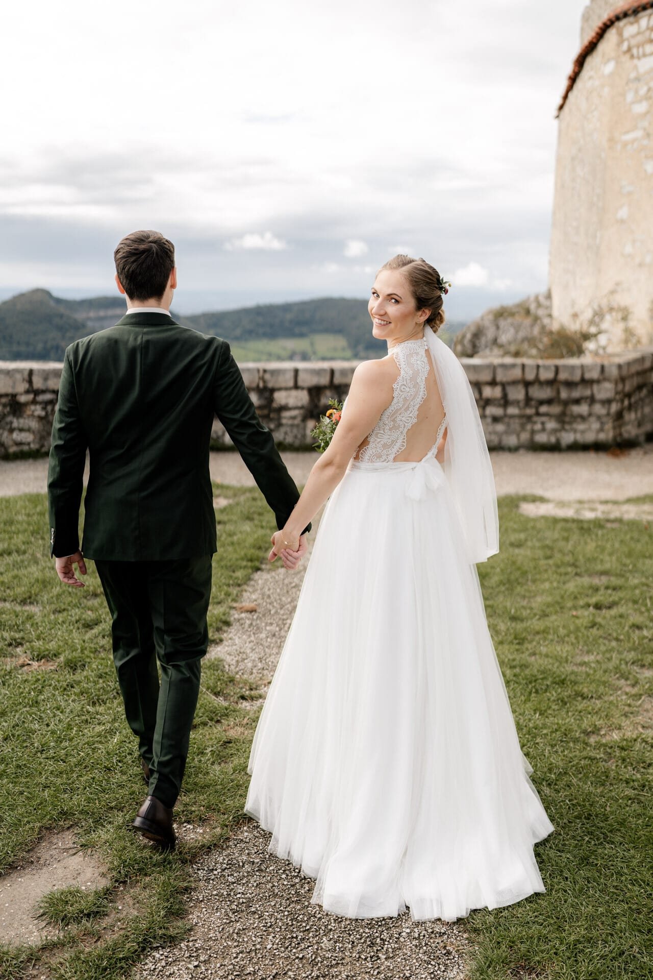 Eine Braut in einem weißen Kleid und ein Bräutigam in einem dunklen Anzug gehen Hand in Hand im Freien auf der Hochzeit Burg Hohenneuffen. Die Braut blickt zurück und lächelt, eingerahmt von einer malerischen Landschaft und einer historischen Steinmauer im Hintergrund.