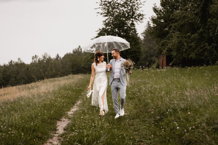 Brautpaar läuft gemeinsam auf Wiese unter Schirm auf ihrer Hochzeit auf Theurerhof Speßhardt