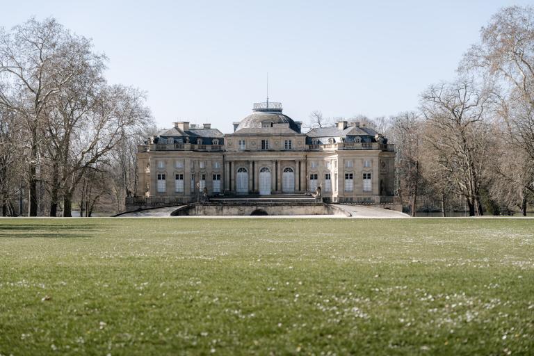 Ein prächtiges historisches Herrenhaus mit Kuppeldach und großen Fenstern steht auf einer erhöhten Steinplattform auf Schloss Monrepos, umgeben von blattlosen Bäumen und einer weitläufigen grünen Wiese - eine zauberhafte Hochzeitskulisse unter klarem Himmel.