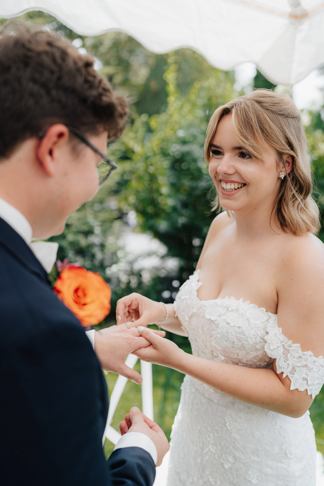 Eine lächelnde Braut in einem schulterfreien Spitzenkleid steckt dem Bräutigam während einer Gewächshaus-Hochzeit im Plangarten Leinfelden-Echterdingen einen Ring an den Finger, mit üppigem Grün im Hintergrund.