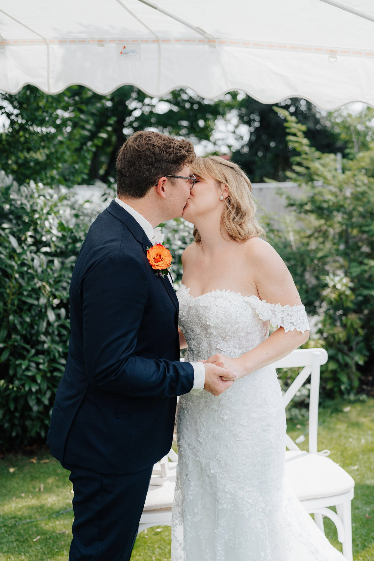 Eine Braut in einem schulterfreien weißen Spitzenkleid und ein Bräutigam in einem marineblauen Anzug mit einer orangefarbenen Boutonniere küssen sich im Freien unter einem weißen Baldachin im Gewächshaus Hochzeit Plangarten Leinfelden-Echterdingen.