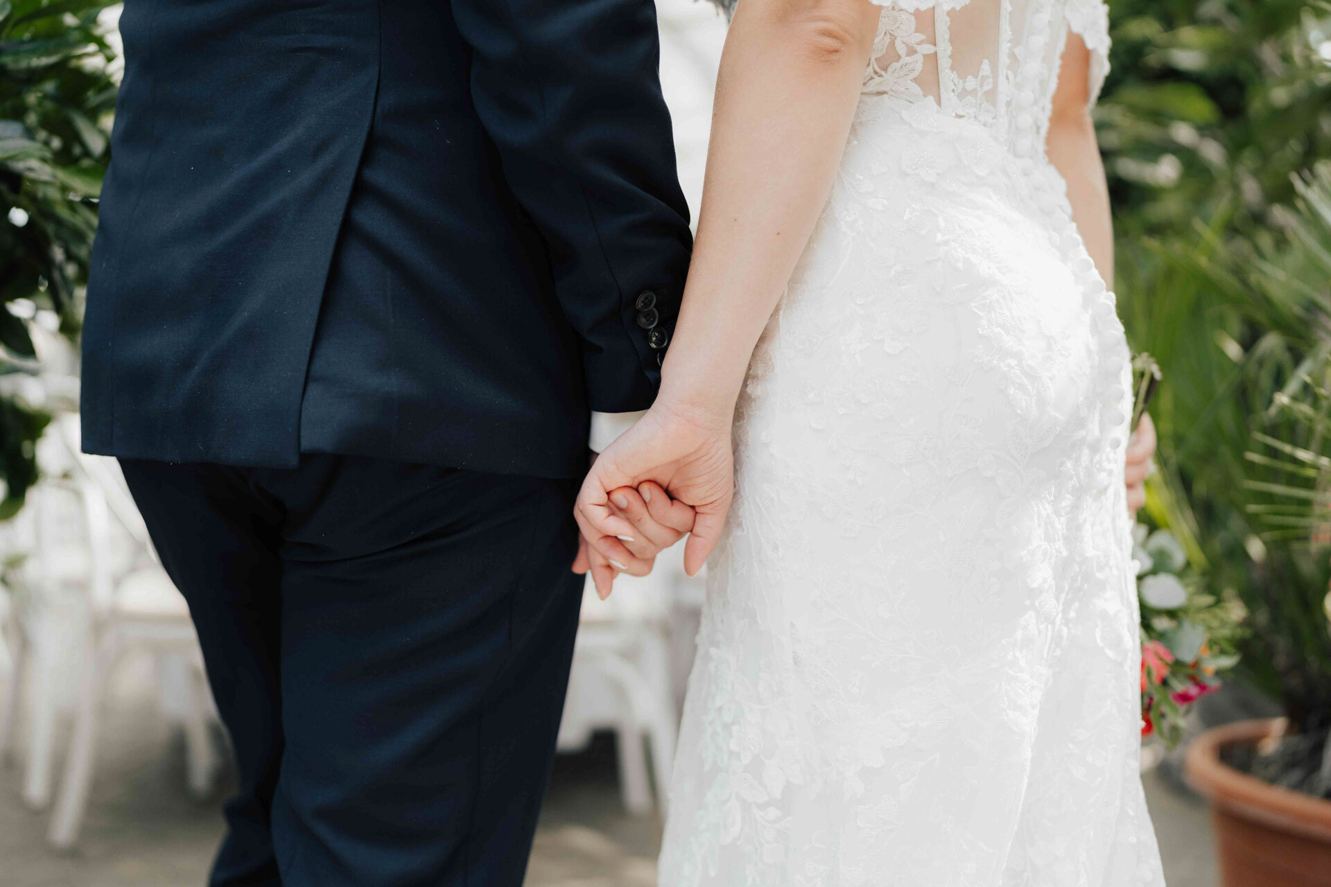 Braut und Bräutigam halten sich an den Händen, fotografiert von hinten bei ihrer Gewächshaus-Hochzeit in Plangarten Leinfelden-Echterdingen. Die Braut trägt ein weißes Spitzenkleid, der Bräutigam einen dunklen Anzug; Grünpflanzen und Stühle füllen den Hintergrund.