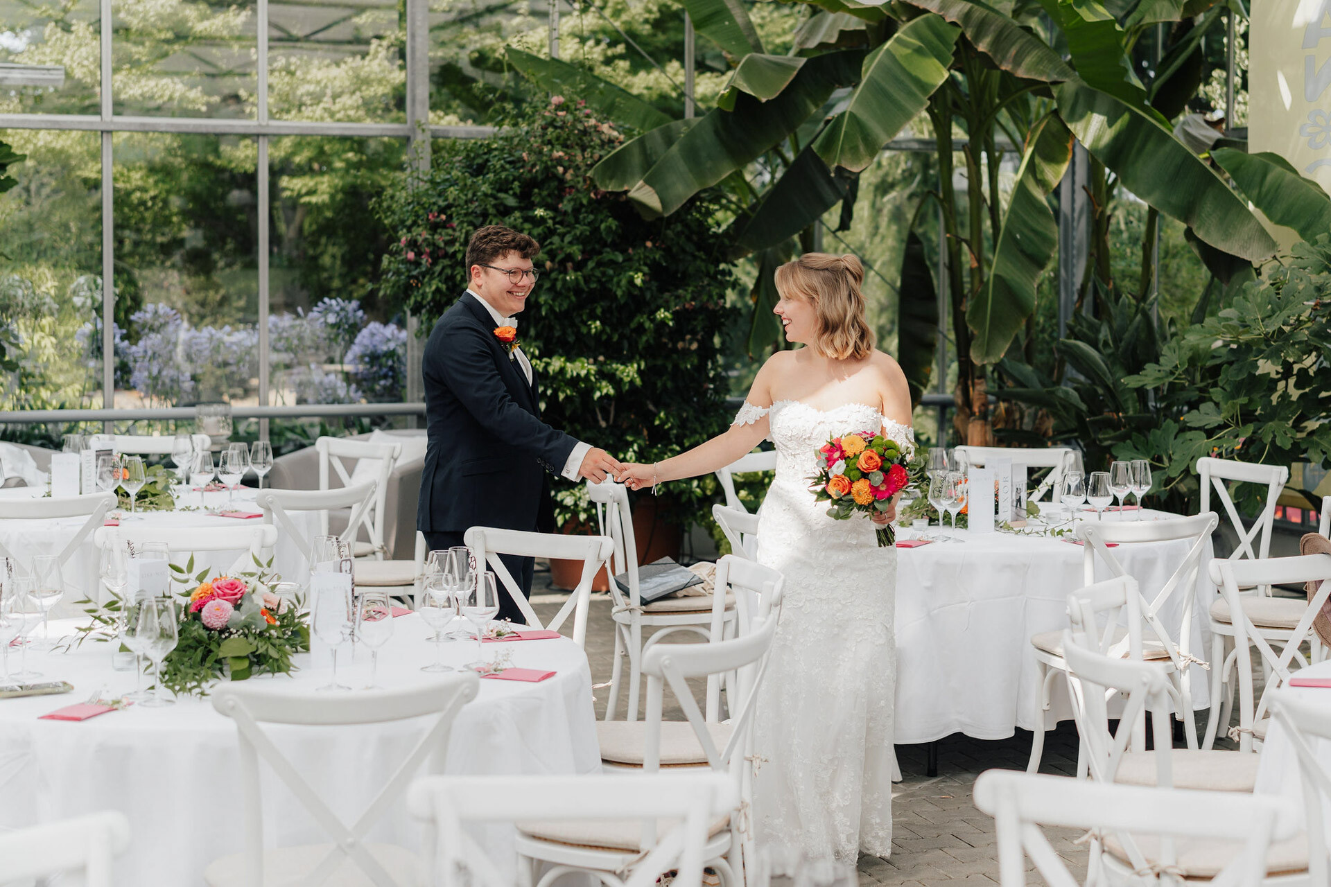Eine Braut und ein Bräutigam lächeln sich an und halten sich an den Händen im hellen, begrünten Gewächshaus Hochzeit Plangarten Leinfelden-Echterdingen, wo runde Tische für einen Hochzeitsempfang gedeckt sind. Die Braut trägt ein weißes Kleid und einen bunten Blumenstrauß.