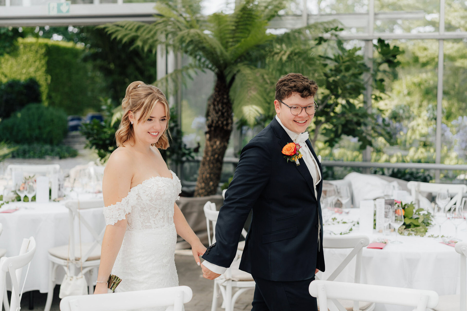 Eine Braut in einem weißen schulterfreien Kleid und ein Bräutigam in einem dunklen Anzug mit einer leuchtend orangefarbenen Boutonniere halten sich an den Händen und lächeln, während sie durch das elegante Gewächshaus Hochzeit im Plangarten Leinfelden-Echterdingen gehen, umgeben von runden Tischen und üppigem Grün.