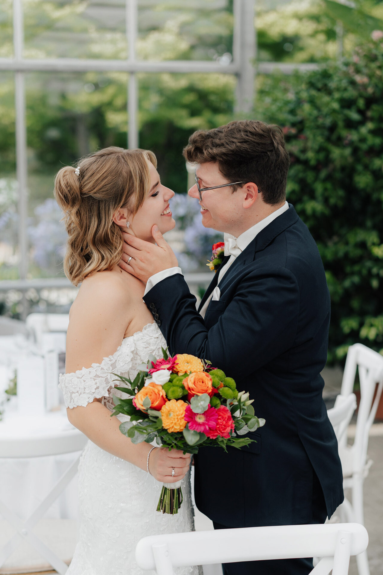 Eine Braut in einem weißen schulterfreien Kleid mit einem bunten Blumenstrauß in der Hand lächelt, während der Bräutigam in einem dunklen Anzug sanft ihr Gesicht berührt. Sie stehen dicht beieinander und schauen sich in die Augen im Gewächshaus Hochzeit Plangarten Leinfelden-Echterdingen.