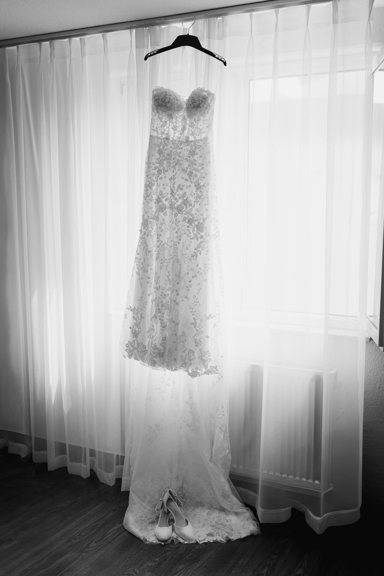 Ein Spitzenhochzeitskleid hängt auf einem schwarzen Bügel vor einem weißen Vorhang, darunter ein Paar weißer Stöckelschuhe - eingefangen in zeitlosem Schwarz und Weiß im Plangarten Leinfelden-Echterdingen.