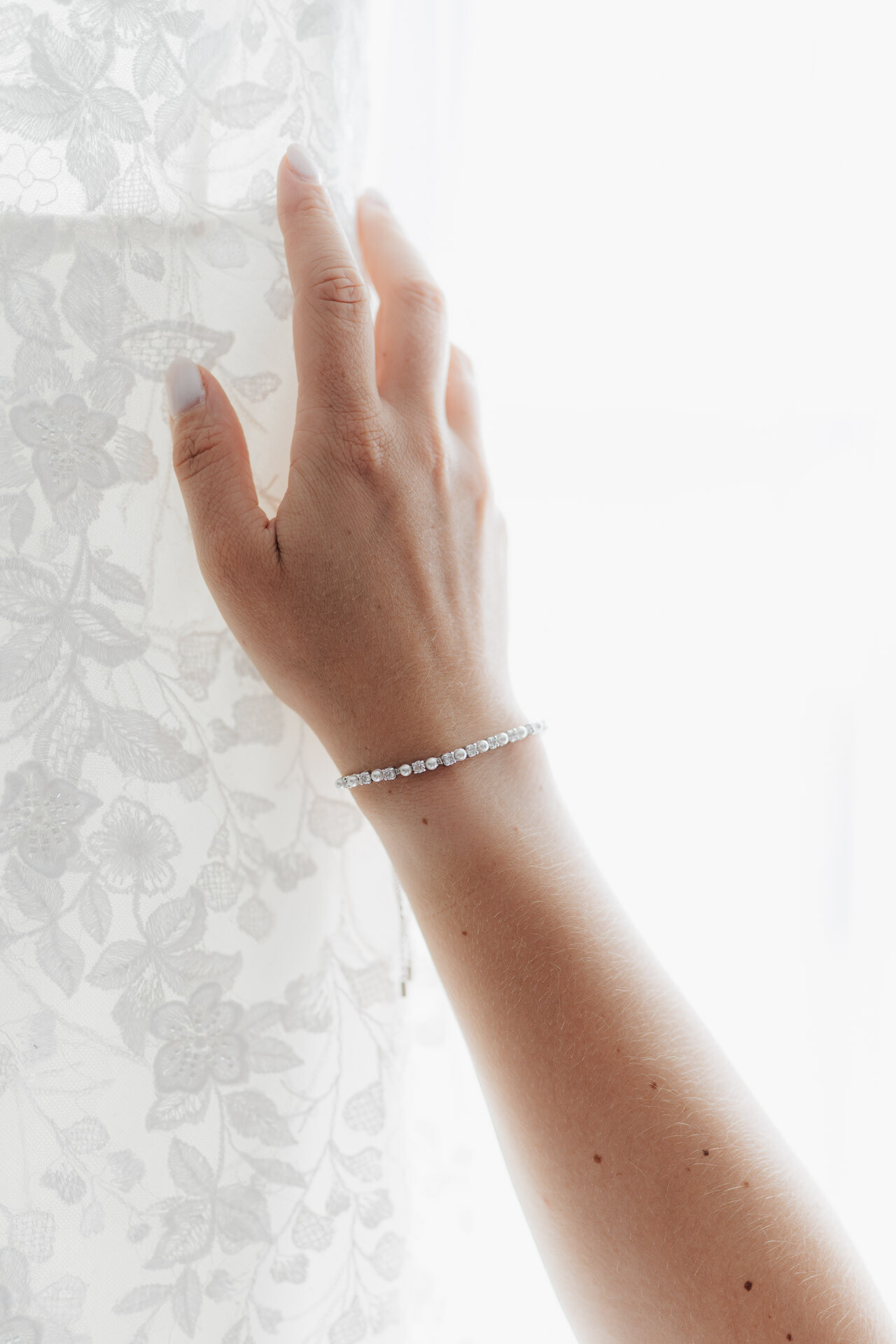 Eine Nahaufnahme einer Hand mit hellem Nagellack, die sanft einen weißen, geblümten Spitzenvorhang berührt. Sie trägt ein zartes Silberarmband mit kleinen Edelsteinen, das die Eleganz einer Gewaechshaus-Hochzeit in Plangarten Leinfelden-Echterdingen perfekt einfängt.