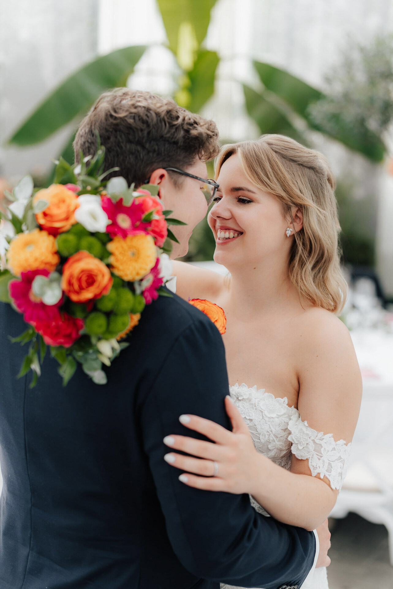 Eine Braut in einem weißen schulterfreien Kleid lächelt einen Bräutigam in einem dunklen Anzug an, der einen bunten Blumenstrauß in der Hand hält. Sie umarmen sich drinnen im Plangarten Leinfelden-Echterdingen und fangen den Charme einer Gewächshaushochzeit mit üppigem Grün im Hintergrund ein.