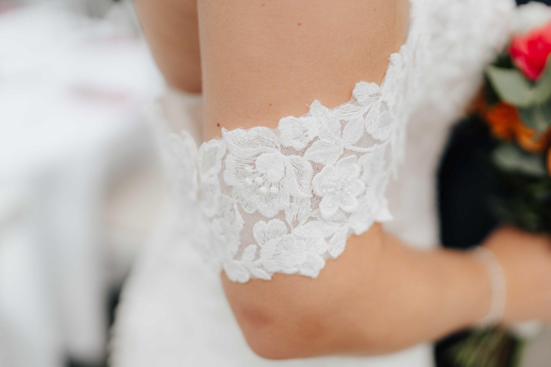 Nahaufnahme des Arms einer Braut in einem weißen Spitzenärmel mit Blumenmustern, die einen Strauß roter und orangefarbener Blumen hält, bei einer Gewächshaushochzeit in Plangarten Leinfelden-Echterdingen, mit einem leicht verschwommenen Hintergrund.