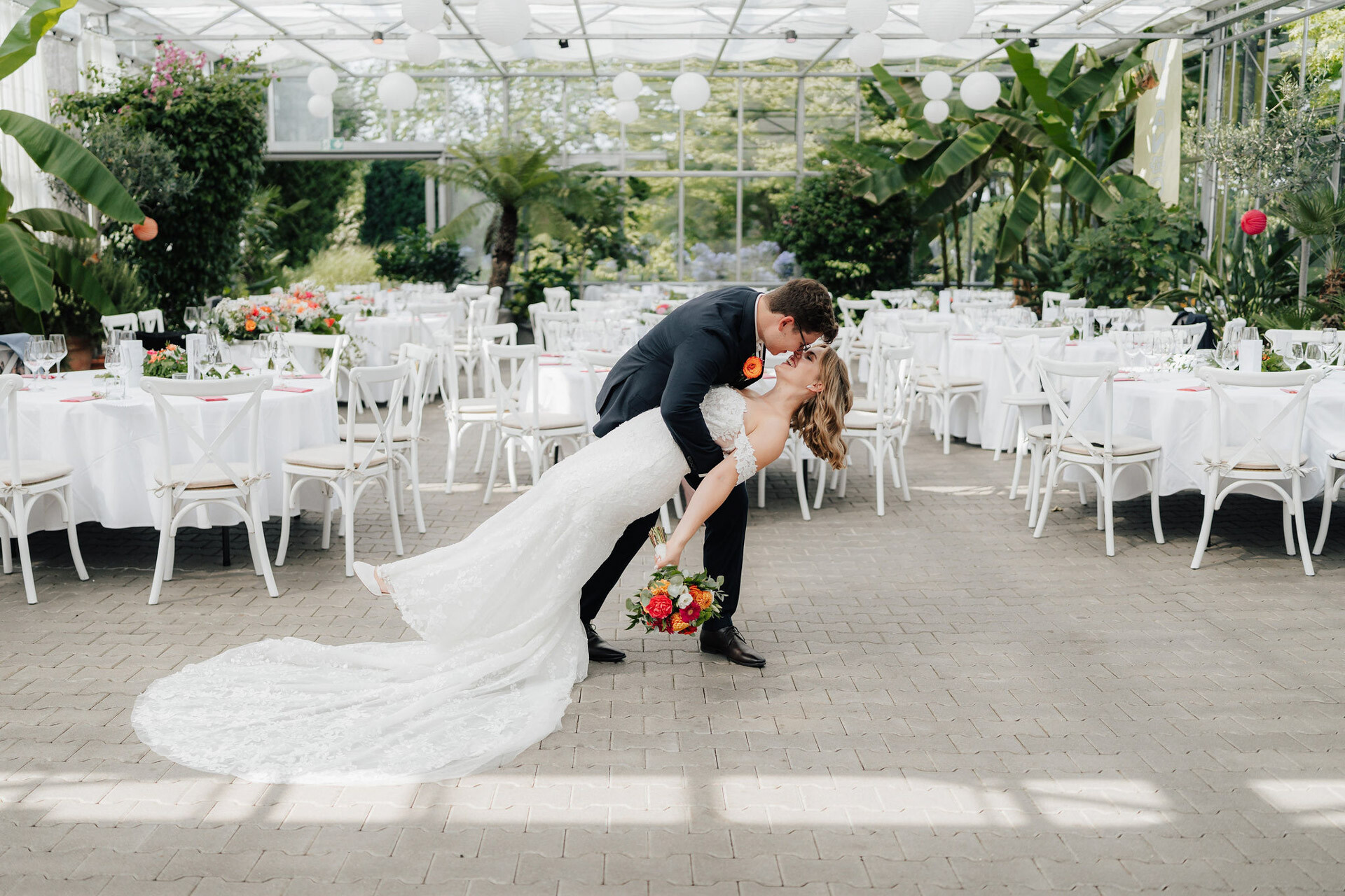 Ein Bräutigam taucht und küsst seine Braut im Gewächshaus Hochzeit im Plangarten Leinfelden-Echterdingen, umgeben von weißen Tischen, Stühlen und hängenden Laternen. Die Braut trägt ein weißes Kleid mit einem Blumenstrauß; der Bräutigam trägt einen schwarzen Anzug.