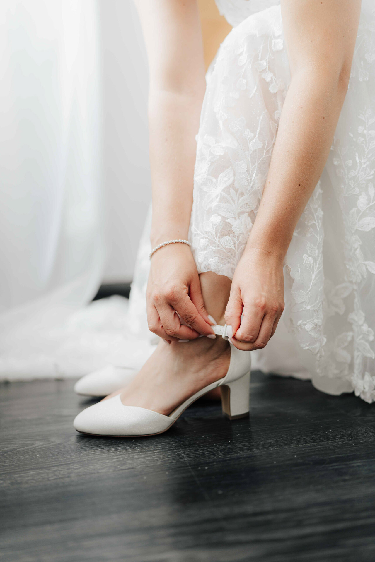 Eine Person in einem weißen Spitzenkleid bückt sich, um den Riemen eines weißen Stöckelschuhs zu schließen. Das Sonnenlicht beleuchtet die Szene im Gewächshaus Hochzeit in Plangarten, Leinfelden-Echterdingen, sanft.