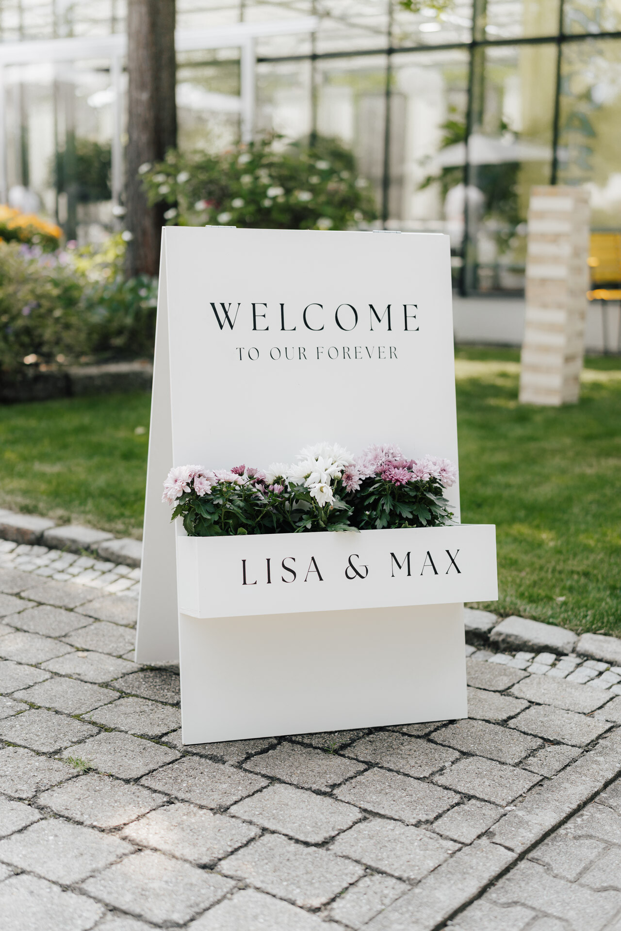 Ein weißes Hochzeitsschild auf einem Steinweg mit der Aufschrift WELCOME TO OUR FOREVER mit LISA & MAX unter einer Schachtel mit rosa und weißen Blumen; im Hintergrund sind Grünpflanzen und das gläserne Gewächshaus im Plangarten Leinfelden-Echterdingen zu sehen.