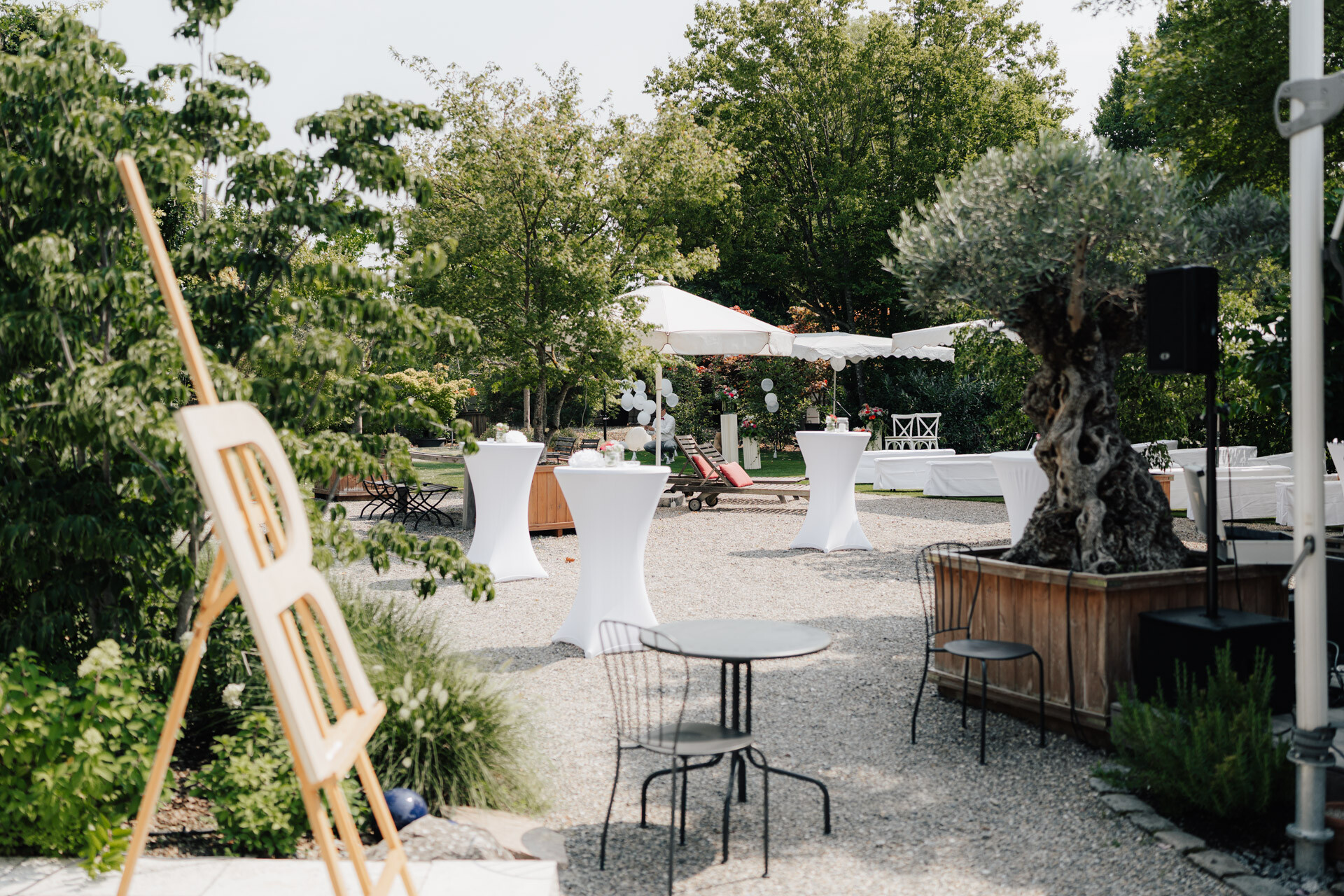 Veranstaltungsraum im Freien im Plangarten Leinfelden-Echterdingen mit Cocktailtischen aus weißem Stoff, Metallstühlen, einer Staffelei und einem Lautsprecher - perfekt für eine entspannte und elegante Garten- oder Gewächshaushochzeit inmitten von üppigem Grün.