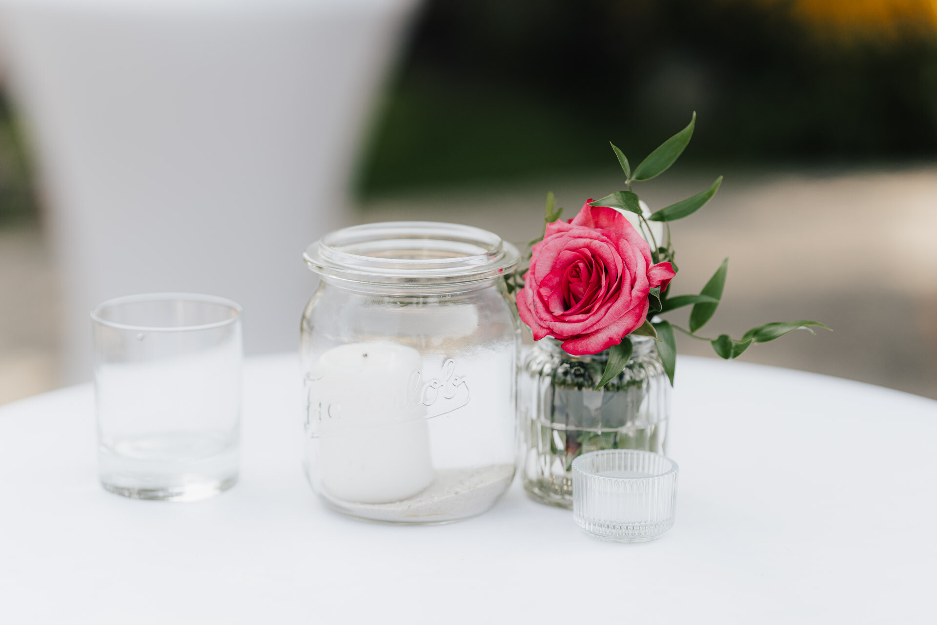Ein kleiner Tisch mit einem gläsernen Votivkerzenhalter, einem Einmachglas mit einer weißen Kerze und einem Glas mit einer rosafarbenen Rose und Grünzeug, der im Gewächshaus Hochzeit Plangarten Leinfelden-Echterdingen im Freien steht.