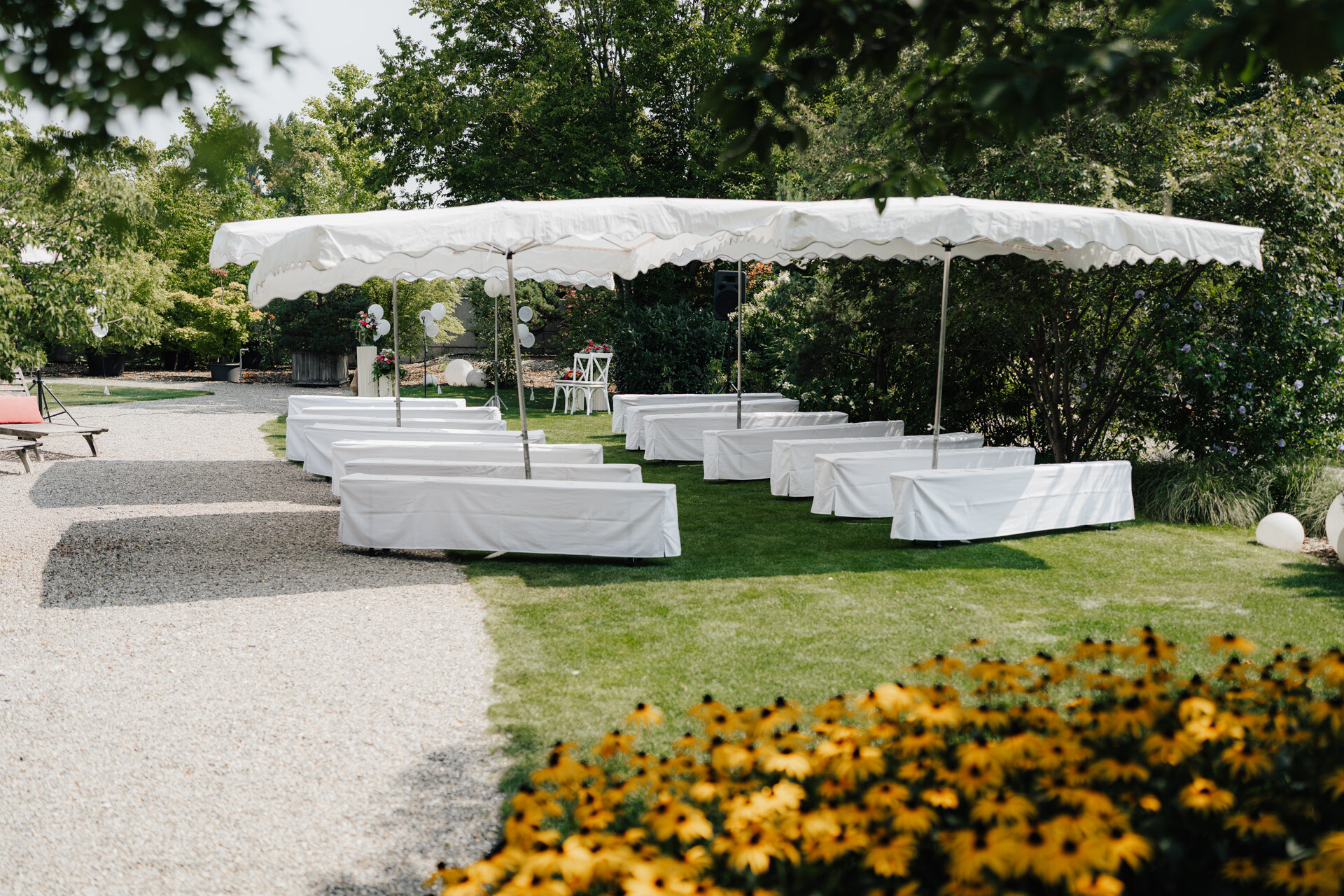 Hochzeitszeremonie im Freien mit weißen Bänken unter einem großen Baldachin im Gewächshaus Hochzeit Plangarten Leinfelden-Echterdingen, umgeben von grünen Bäumen, gelben Blumen und einem nahegelegenen Kiesweg auf der Wiese.