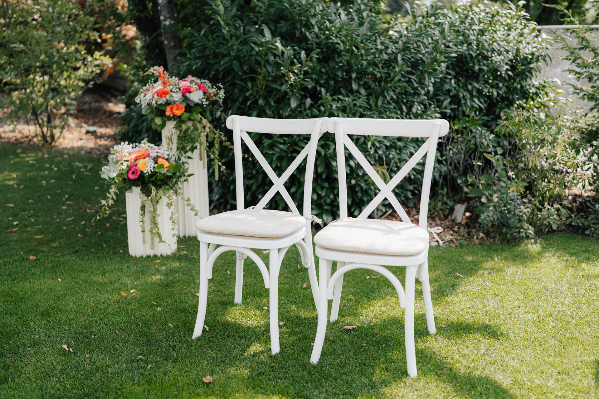 Zwei weiße Holzstühle mit Kissen stehen auf grünem Gras neben weißen Kisten mit bunten Blumenarrangements. Sträucher und Bäume bilden eine grüne Kulisse im Plangarten des Gewaechshauses Hochzeit in Leinfelden-Echterdingen.