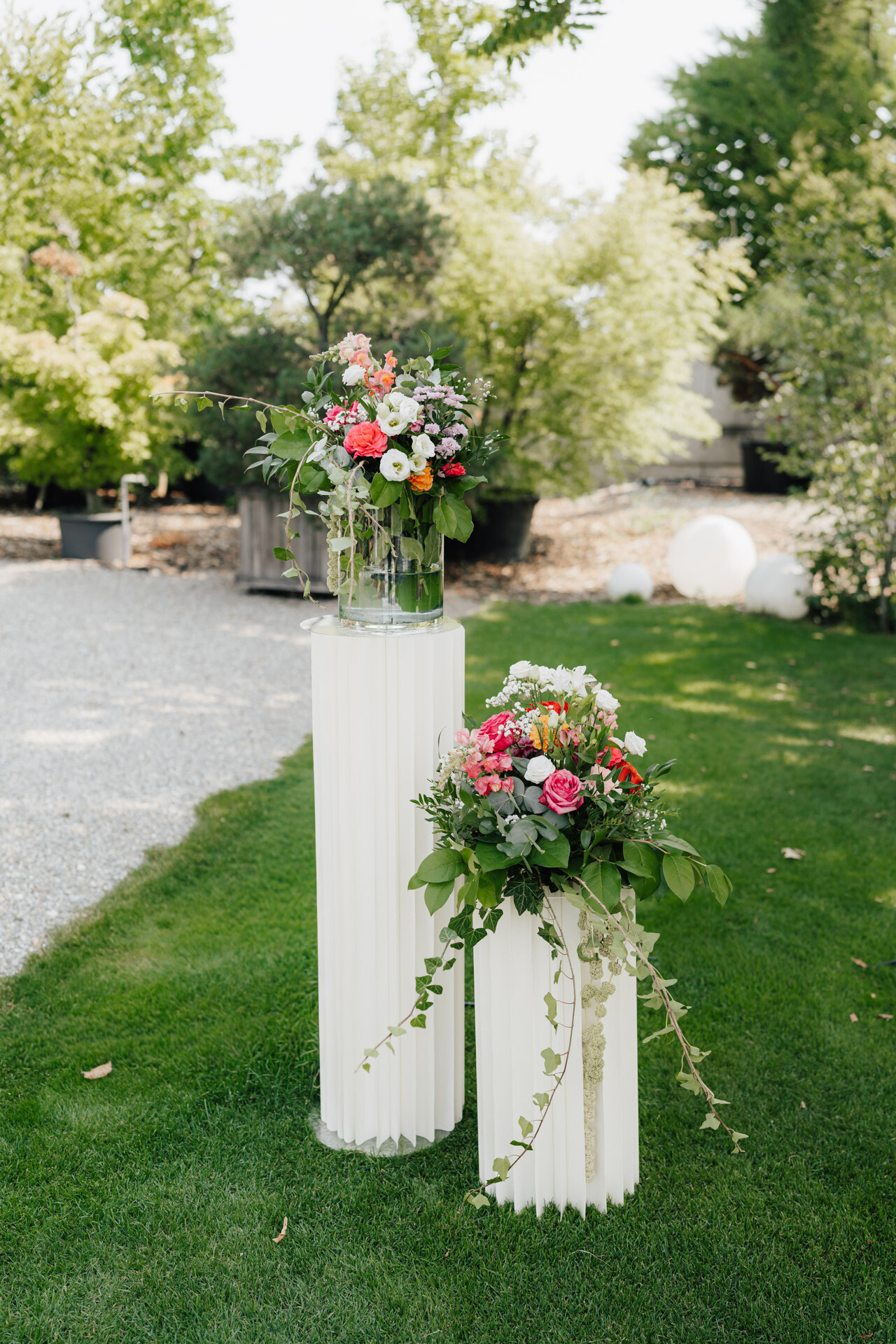 Auf zwei weißen Sockeln auf Gras stehen Vasen mit farbenfrohen Blumenarrangements, darunter rosa, rote, gelbe und weiße Blüten mit Grünzeug - perfekt für eine Gewächshaus-Hochzeit im Plangarten Leinfelden-Echterdingen. Bäume und Sträucher bilden einen üppigen Gartenhintergrund.