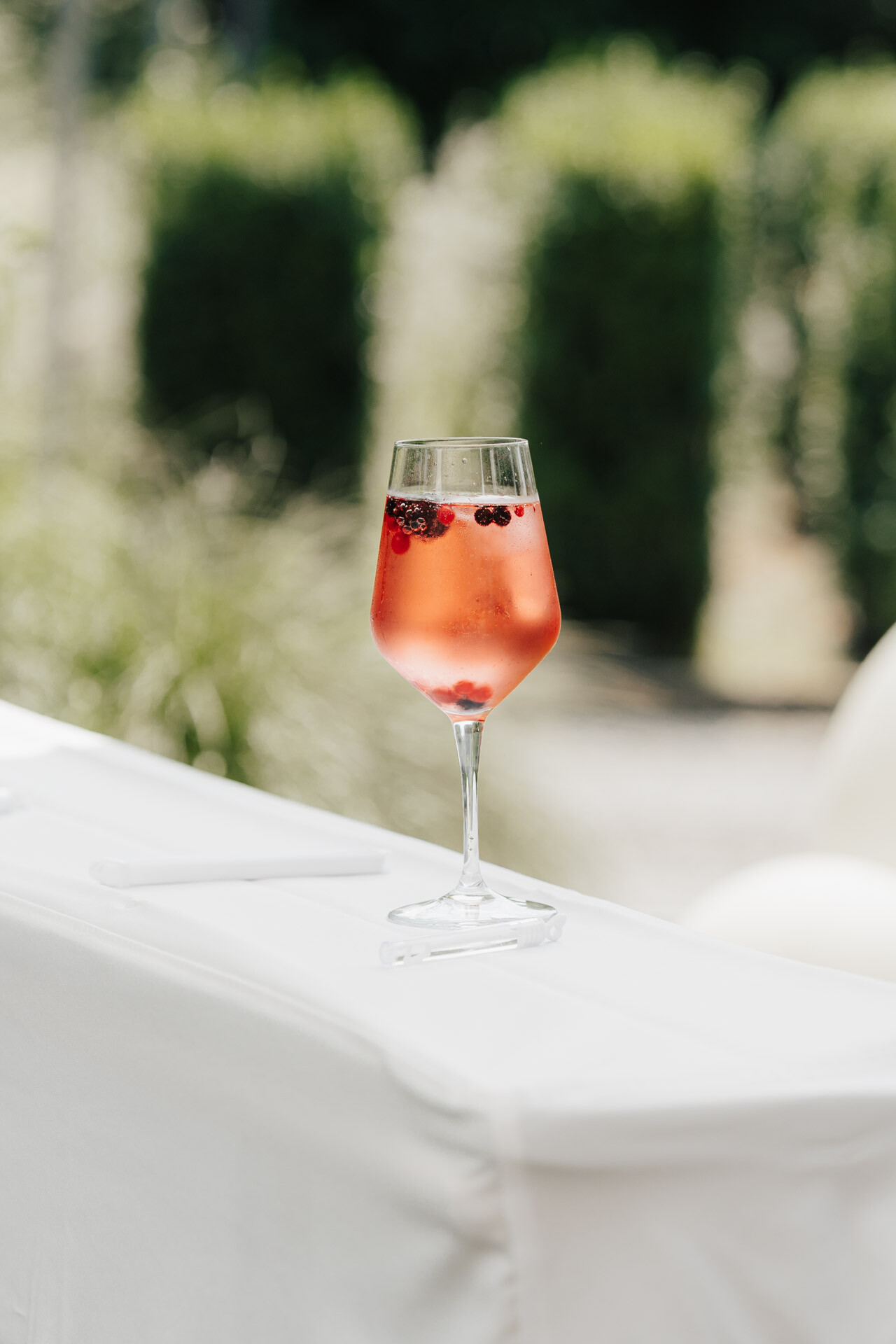 Ein mit rosa Sekt und Beeren gefülltes Weinglas steht auf einem weißen Tischtuch im Freien bei einer Gewächshaus-Hochzeit, mit grünem Laub im Hintergrund in Plangarten Leinfelden-Echterdingen.