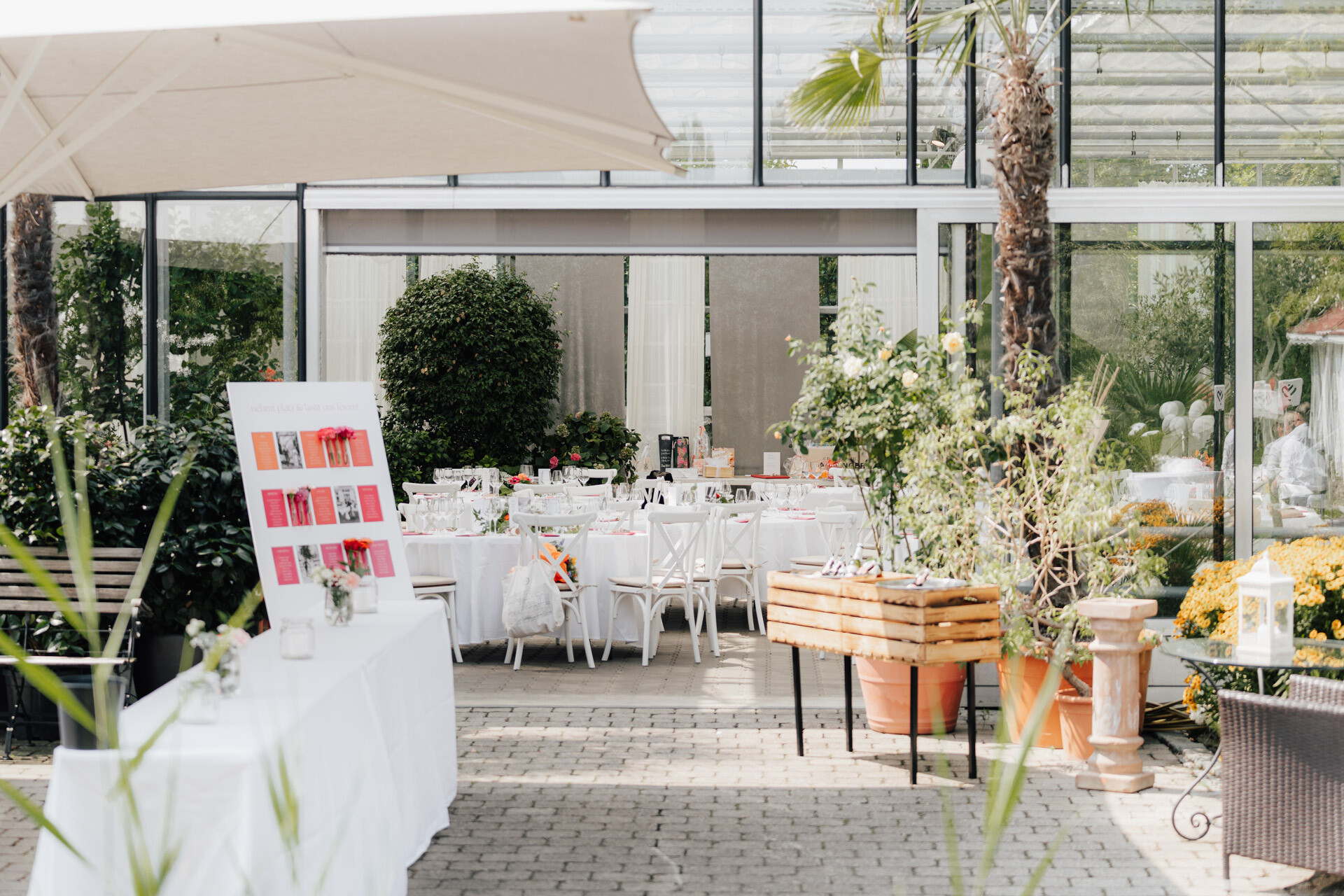 Der Plangarten Leinfelden-Echterdingen ist ein heller, eleganter Veranstaltungsort im Freien mit weiß eingedeckten Tischen, dekorativen Pflanzen und einer Anzeigetafel unter einem großen Schirm neben einem modernen Gewächshaus.