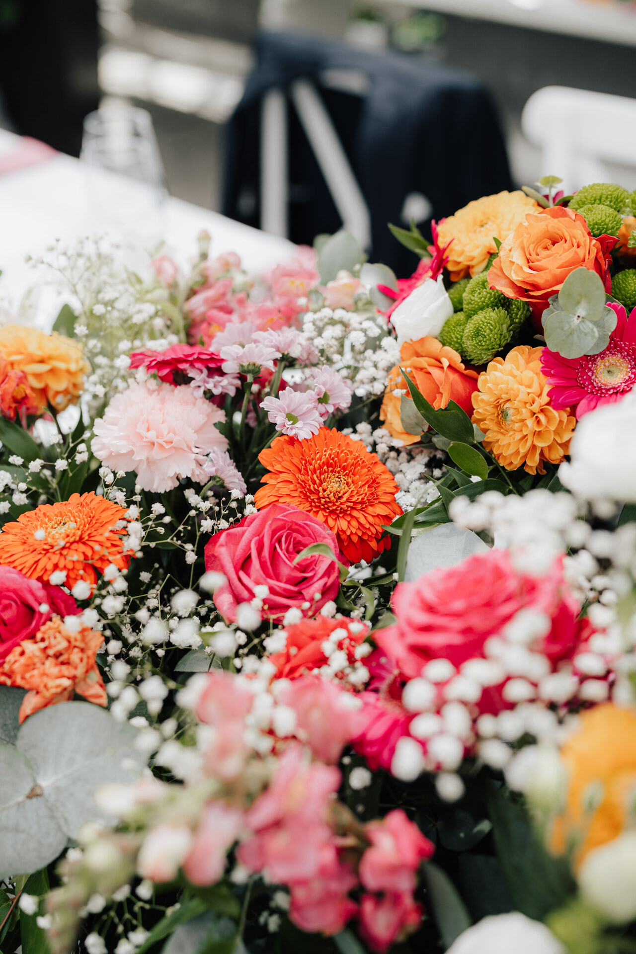 Ein farbenfrohes Blumenarrangement mit orangefarbenen Ringelblumen, rosafarbenen Rosen und Eukalyptusblättern bereichert jede Gewaechshaus-Hochzeit im Plangarten Leinfelden-Echterdingen, das elegant auf einem Tisch mit verschwommenem Hintergrund präsentiert wird.