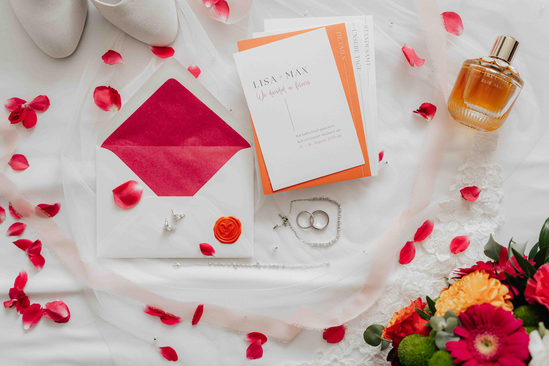 Eine flache Anordnung von Hochzeitsartikeln: ein offener Umschlag mit rotem Futter, eine Einladung für Lisa und Max, Schmuck, Parfüm, weiße Schuhe und verstreute rote Rosenblüten mit einem Strauß - die perfekte Inspiration für eine Gewächshaus-Hochzeit im Plangarten Leinfelden-Echterdingen.
