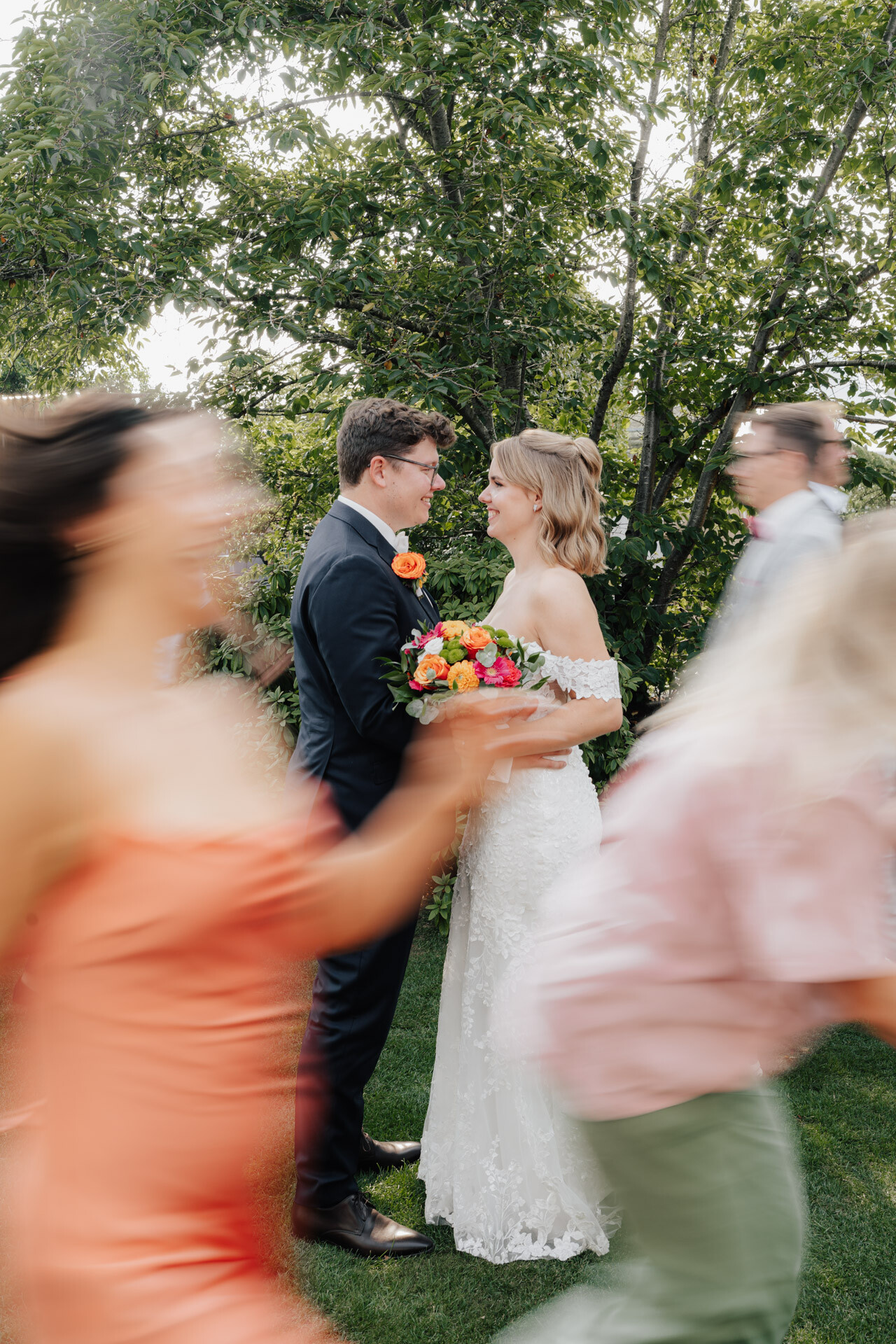 Eine Braut und ein Bräutigam stehen lächelnd im Freien im Plangarten Leinfelden-Echterdingen und halten einen bunten Blumenstrauß. Gäste in farbenfroher Kleidung ziehen vorbei und schaffen eine lebendige, fröhliche Gewächshaus-Hochzeitsatmosphäre inmitten üppig grüner Bäume im Hintergrund.