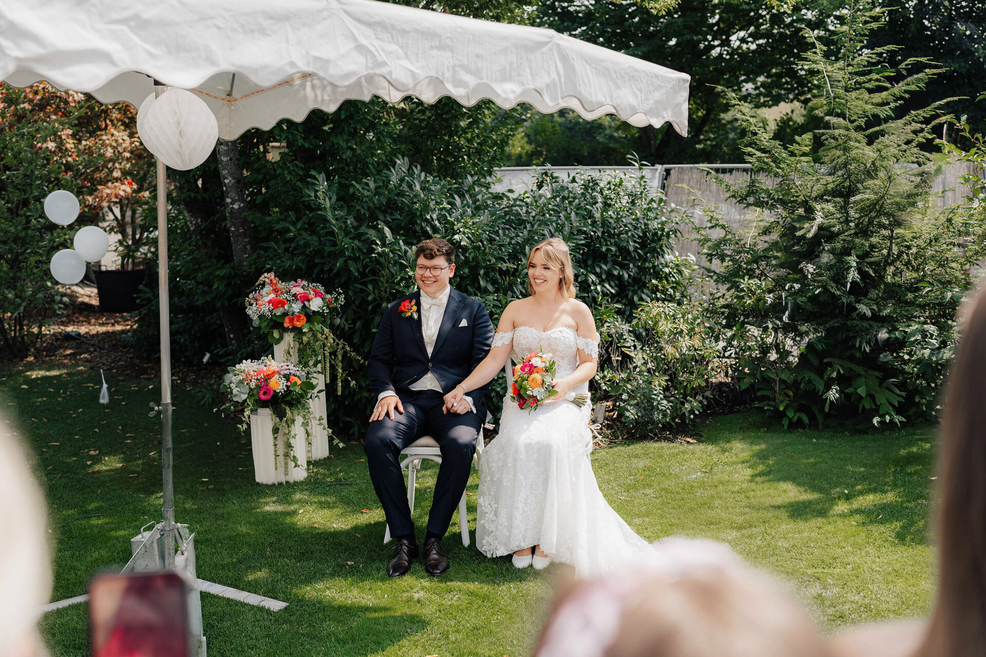 Ein Mann und eine Frau sitzen auf einem Stuhl unter einem weißen Zelt bei einer Gewächshaushochzeit in Plangarten, Leinfelden-Echterdingen.