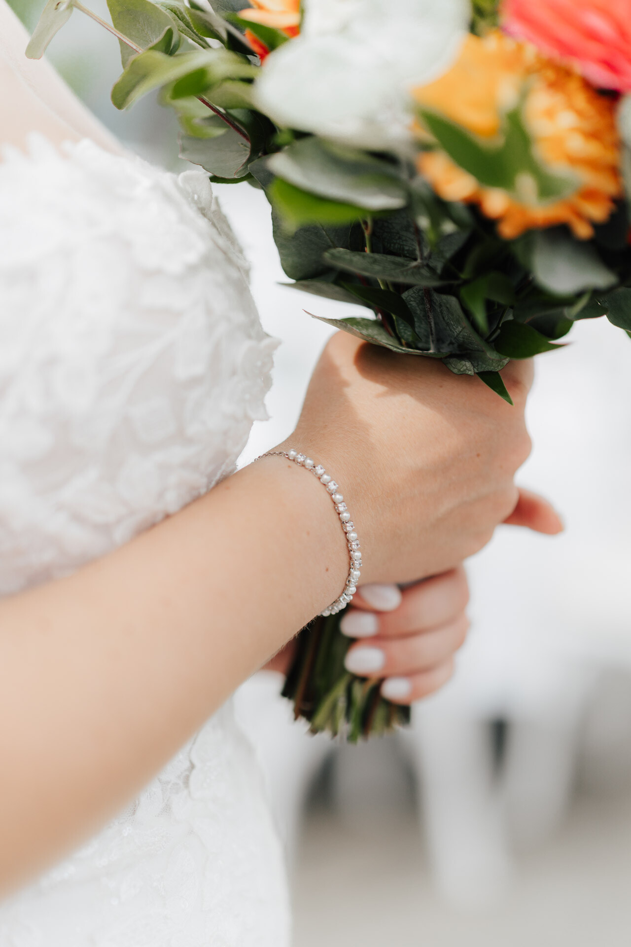 Eine Braut in einem Spitzenhochzeitskleid hält einen bunten Blumenstrauß im Gewächshaus Hochzeit Plangarten Leinfelden-Echterdingen. Sie trägt ein zartes Diamantarmband, und ihre manikürten, weiß lackierten Nägel sind auf dem Bild hervorgehoben.