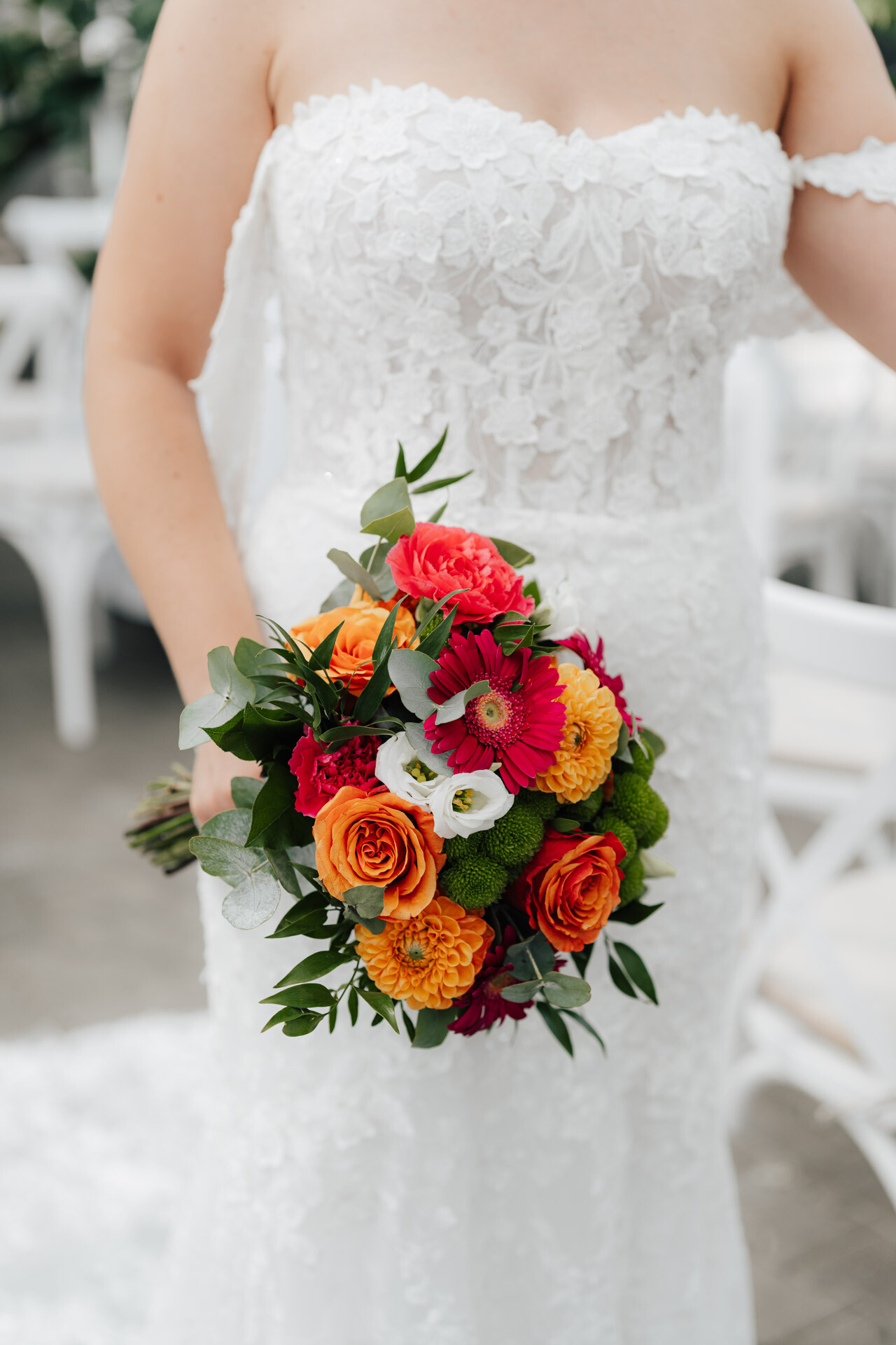 Eine Braut in einem schulterfreien Hochzeitskleid aus weißer Spitze hält einen leuchtenden Strauß aus orangefarbenen, roten und weißen Blumen im Gewächshaus Hochzeit Plangarten Leinfelden-Echterdingen. Ihr Gesicht ist nicht zu sehen. Im Hintergrund sind weiße Stühle zu sehen.