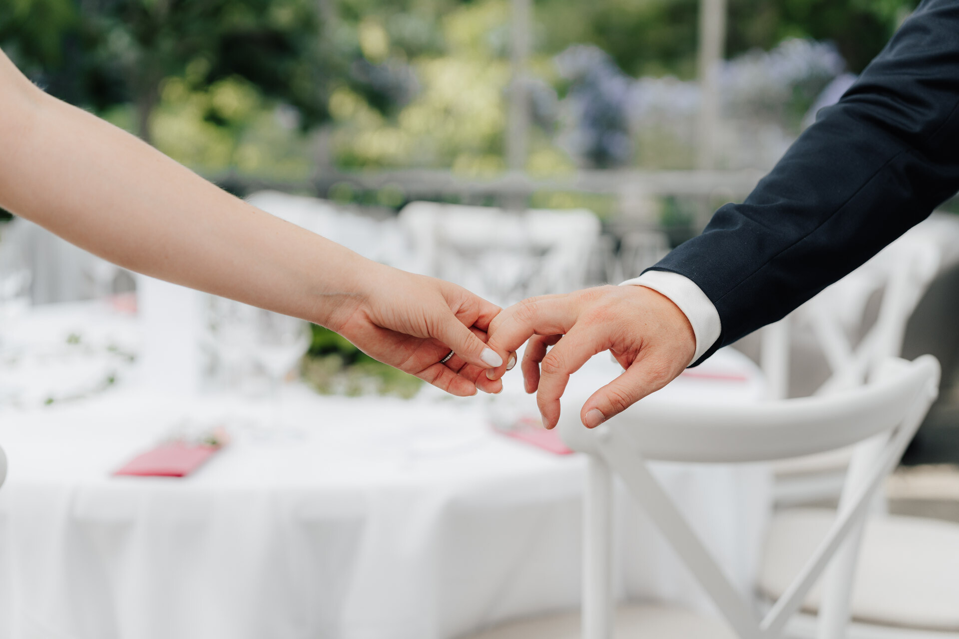 Eine Nahaufnahme von zwei Menschen, die sich sanft an den Händen halten, einer in einem weißen Kleid und der andere in einem dunklen Anzug, auf einer Gewächshaus-Hochzeit. Ein geschmückter Tisch mit weißen Tischdecken und Stühlen im Plangarten Leinfelden-Echterdingen ist im Hintergrund verschwommen.