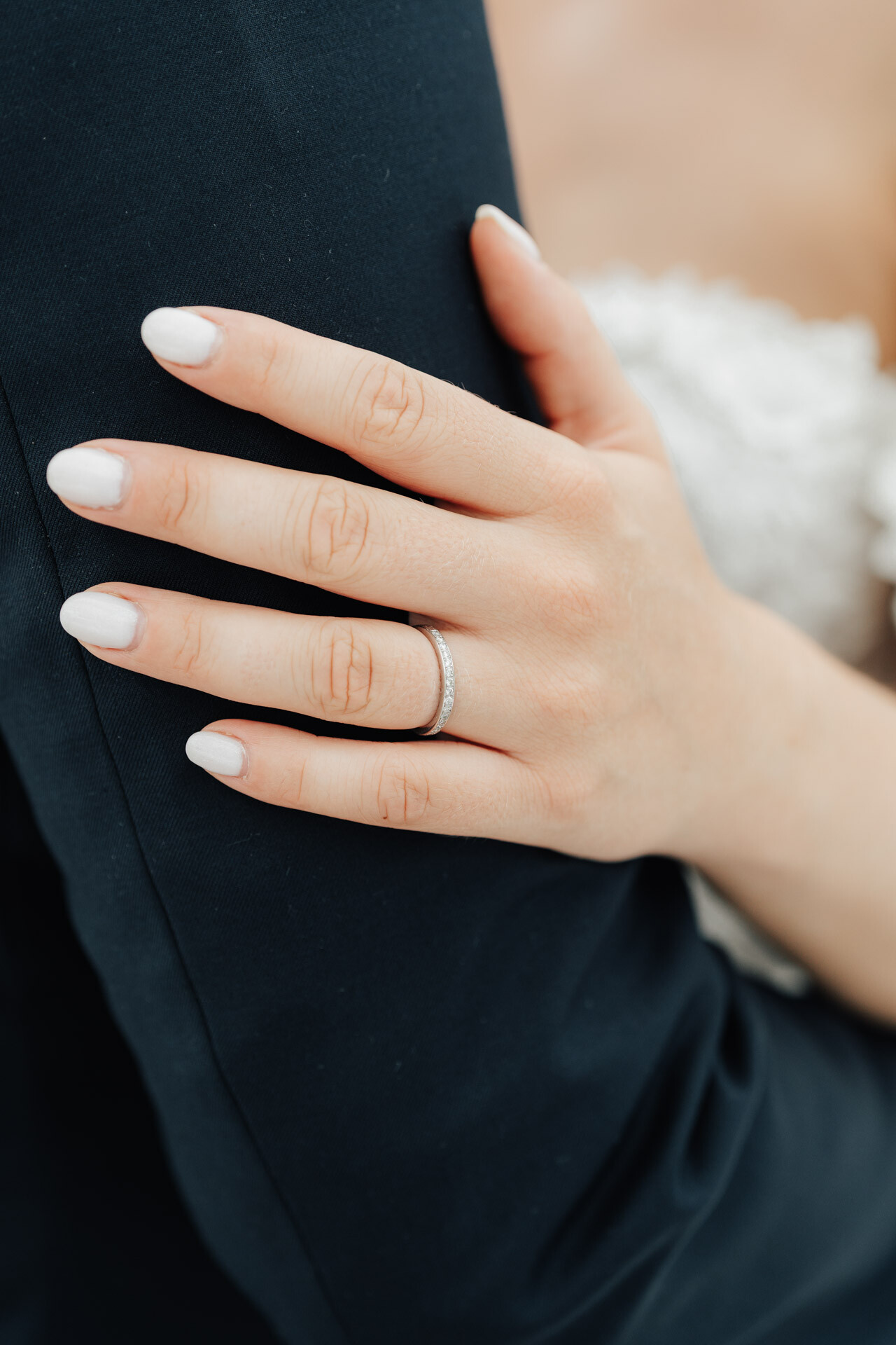 Eine Nahaufnahme einer Frauenhand mit französischer Maniküre und Diamant-Hochzeitsring ruht auf dem Arm eines Mannes in einem dunklen Anzug und fängt einen eleganten Moment auf einer Gewächshaus-Hochzeit in Plangarten Leinfelden-Echterdingen ein.