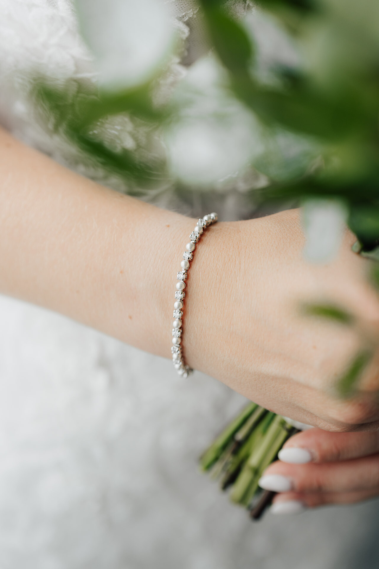 Nahaufnahme einer Frauenhand, die einen Strauß grüner Stiele im Gewächshaus Hochzeit Plangarten Leinfelden-Echterdingen hält. Sie trägt ein zartes Diamant-Tennisarmband und ein weißes Spitzenkleid. Ihre Nägel sind in einem zarten Weißton manikürt.
