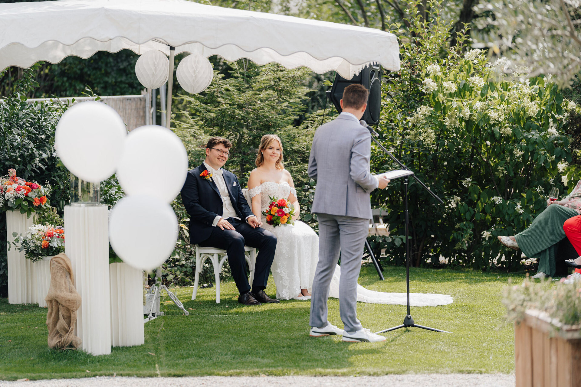 Braut und Bräutigam sitzen bei ihrer Gewächshaus-Hochzeit in Plangarten Leinfelden-Echterdingen unter einem weißen Baldachin und lauschen der Rede eines Mannes im grauen Anzug. Weiße Luftballons und Blumenarrangements schmücken die üppige Gartenanlage.
