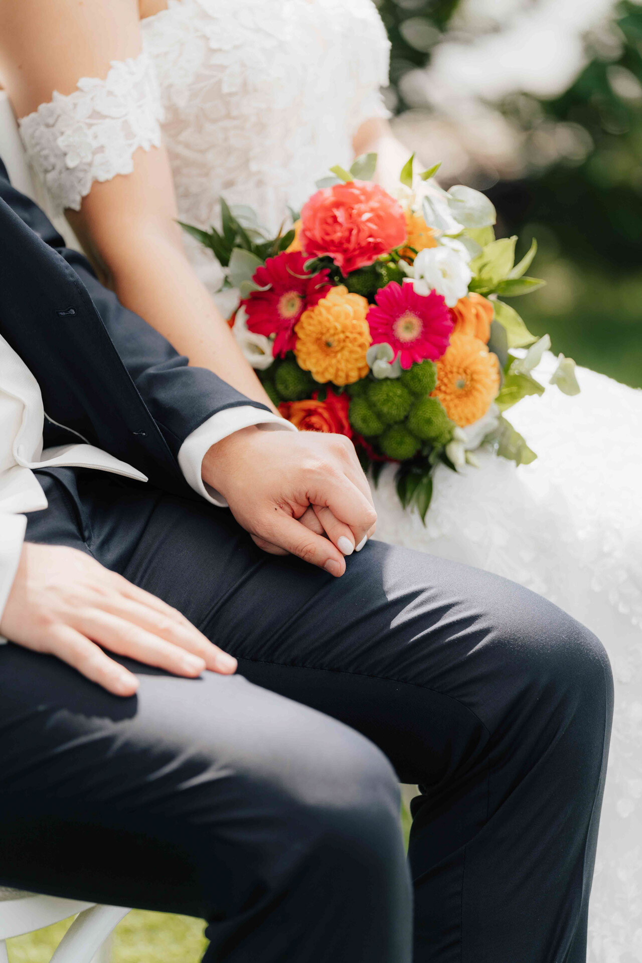 Eine Braut und ein Bräutigam sitzen eng beieinander und halten sich an den Händen im Gewächshaus Hochzeit in Plangarten Leinfelden-Echterdingen. Das weiße Spitzenkleid und der Strauß der Braut verleihen ihr Charme, während der dunkle Anzug des Bräutigams die romantische Umgebung ergänzt.