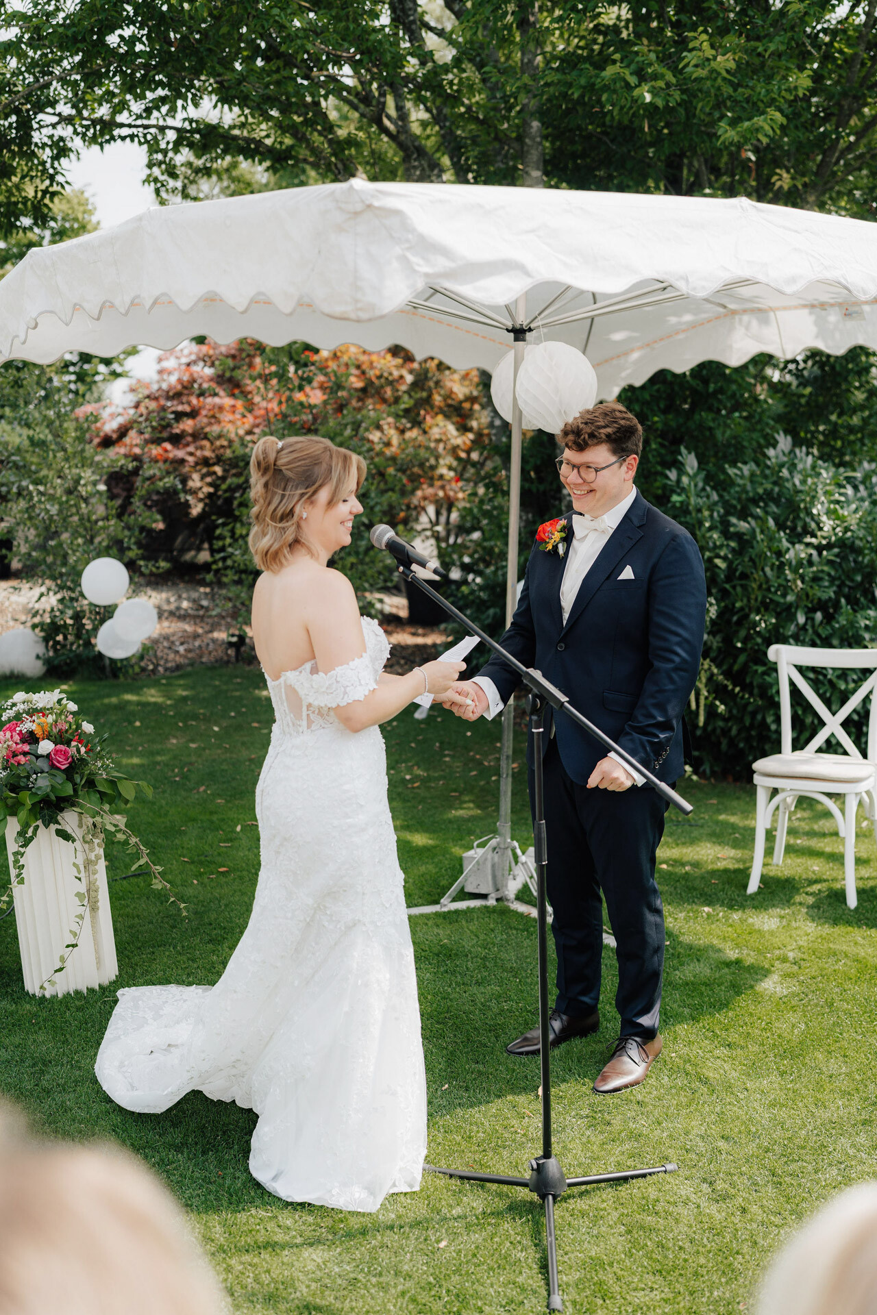 Eine Braut in einem weißen schulterfreien Kleid und ein Bräutigam in einem dunklen Anzug stehen lächelnd unter einem weißen Baldachin im Gewächshaus Hochzeit Plangarten Leinfelden-Echterdingen und tauschen während ihrer Hochzeitszeremonie im Freien die Ringe aus.