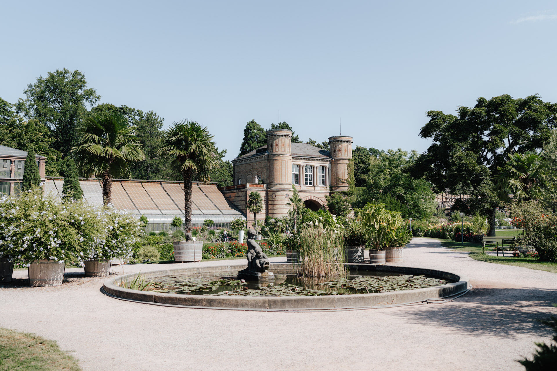 Ein runder Teich mit einer kleinen Statue in der Mitte, umgeben von üppigen Pflanzen, Palmen und Blumen, bildet eine romantische Kulisse für eine Hochzeit bei Tante Polly Karlsruhe, mit einem historischen Backsteingebäude und Gewächshäusern im Hintergrund unter einem klaren blauen Himmel.