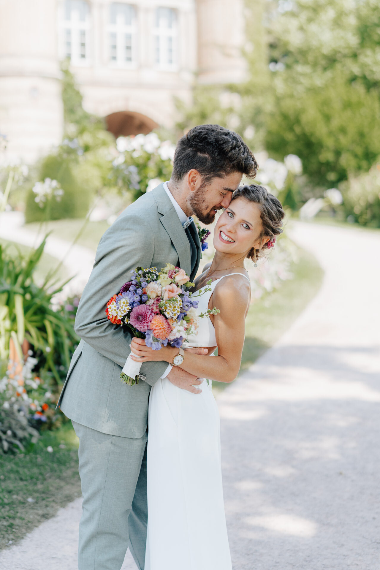 Ein Bräutigam in einem hellgrauen Anzug umarmt und küsst die Stirn seiner lächelnden Braut in einem weißen Kleid, die einen bunten Blumenstrauß in der Hand hält, im Freien an einem sonnigen Tag bei Hochzeit Tante Polly Karlsruhe, umgeben von Grün und Blumen.