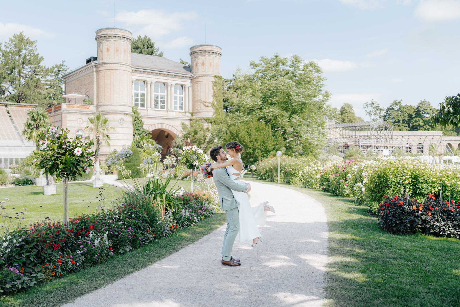 Ein Bräutigam hebt seine Braut freudig auf einen Gartenweg bei Hochzeit Tante Polly Karlsruhe, umgeben von bunten Blumen, Grün und einem historischen Gebäude unter einem strahlenden, sonnigen Himmel.