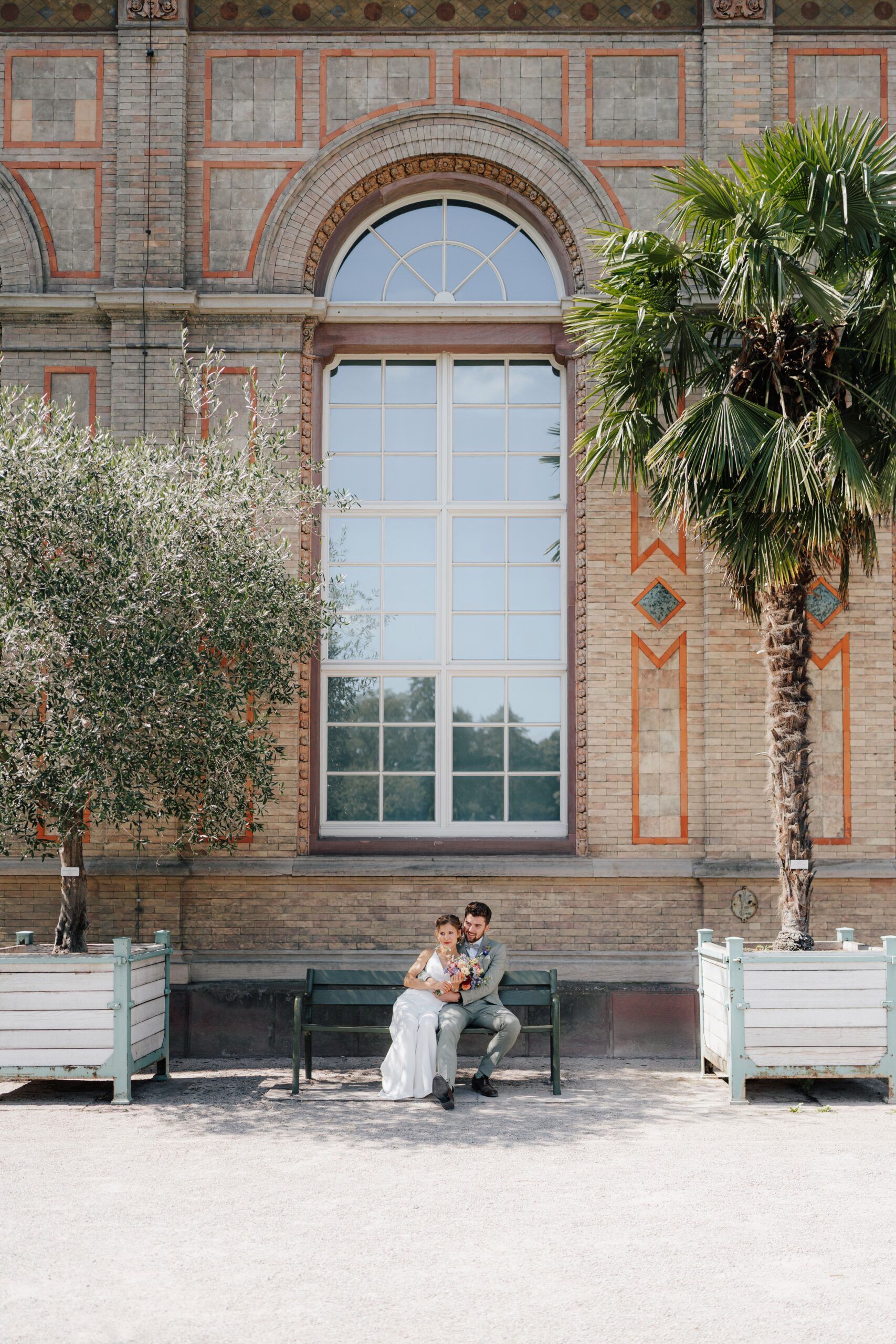 Ein Paar in Hochzeitskleidung sitzt auf einer Bank vor einem großen Fenster bei Hochzeit Tante Polly Karlsruhe, umgeben von zwei Topfbäumen - einer Palme und einer Olive. Die helle Szene im Freien unterstreicht den Charme des Backsteingebäudes.