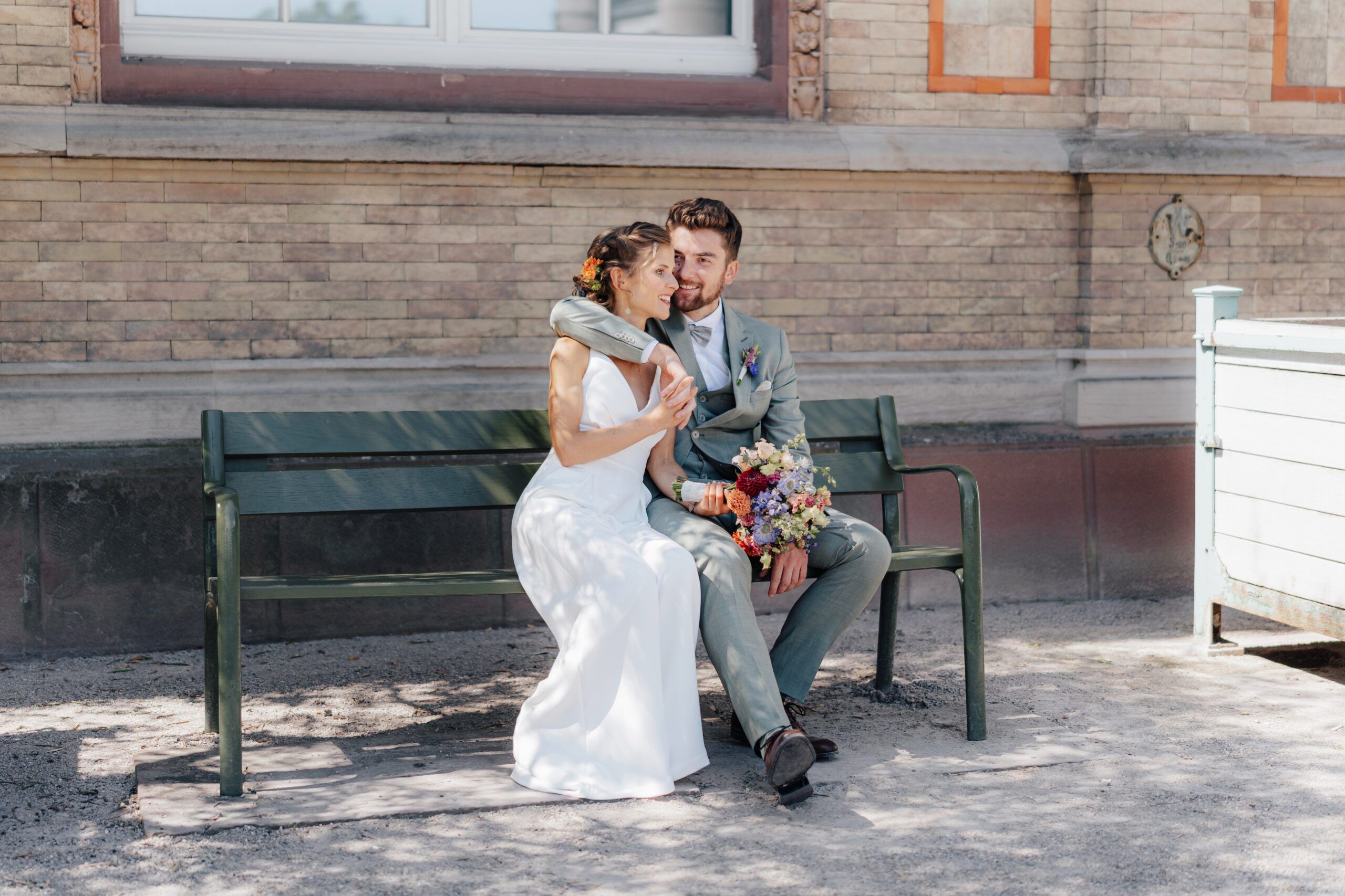 Ein frisch vermähltes Paar sitzt eng beieinander auf einer grünen Bank im Freien bei Hochzeit Tante Polly Karlsruhe. Die Braut in einem weißen Kleid lächelt den Bräutigam mit einem bunten Blumenstrauß an. Vor der Kulisse eines Backsteingebäudes sehen sie glücklich und liebevoll aus.
