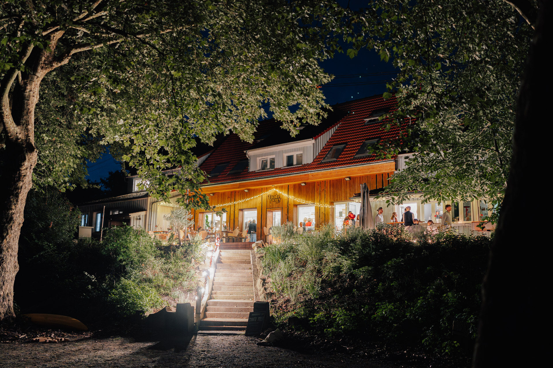 Ein warm beleuchtetes Haus mit rotem Dach und Holzfassade ist nachts von Bäumen umgeben. Bei Hochzeit Tante Polly Karlsruhe säumen Lichter die Treppe hinauf zur Terrasse, auf der sich die Menschen versammeln, und schaffen eine einladende Atmosphäre.