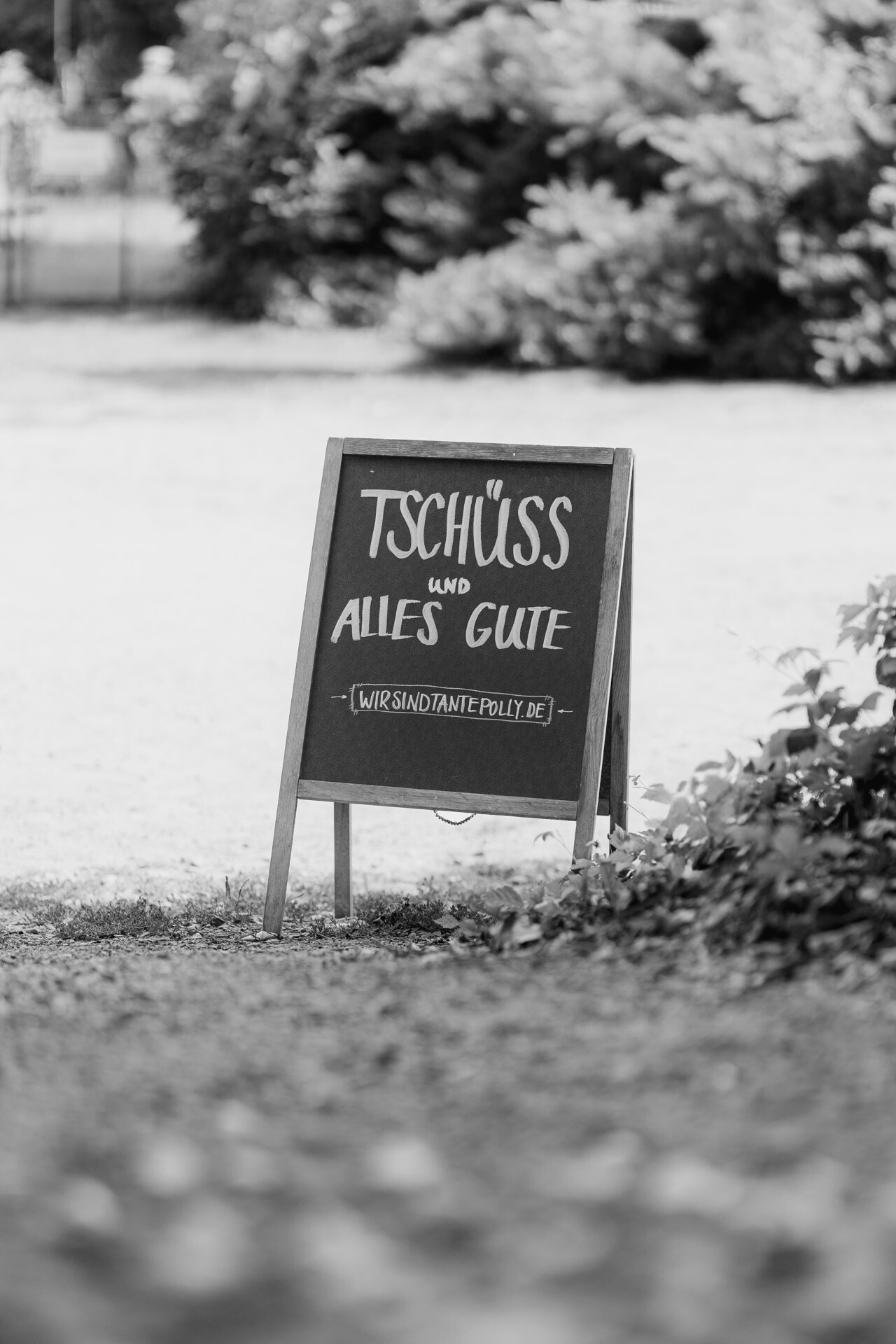 Ein Kreideschild im Freien mit der Aufschrift Tschüss und alles Gute und [WIR SIND TANTE POLLY.DE] in Deutsch, umgeben von Gras und grünen Büschen. Perfekt für eine Hochzeit bei Tante Polly Karlsruhe. Das schwarz-weiße Bild hat verschwommene Bäume im Hintergrund.