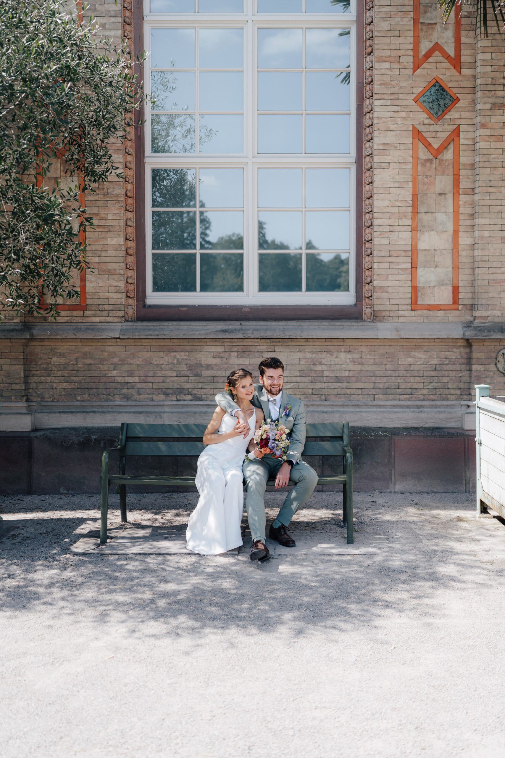 Eine Braut und ein Bräutigam sitzen dicht gedrängt auf einer grünen Bank vor der Hochzeit Tante Polly Karlsruhe, halten einen bunten Blumenstrauß und lächeln an einem sonnigen Tag vor dem großen Backsteingebäude mit hohen Fenstern.
