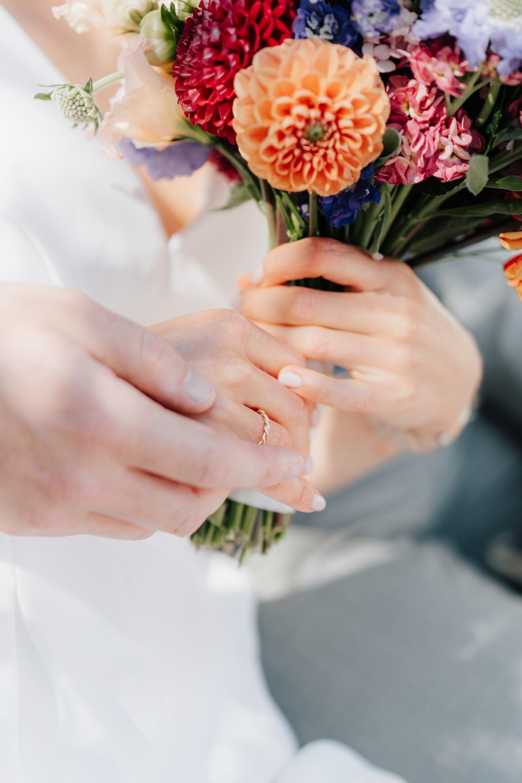 Eine Nahaufnahme von zwei Menschen, die sich an den Händen halten, wobei der eine einen goldenen Ring trägt, während er einen bunten Blumenstrauß mit orangefarbenen, roten und violetten Blüten in der Hand hält, um einen romantischen Hochzeitsmoment bei Tante Polly in Karlsruhe einzufangen.