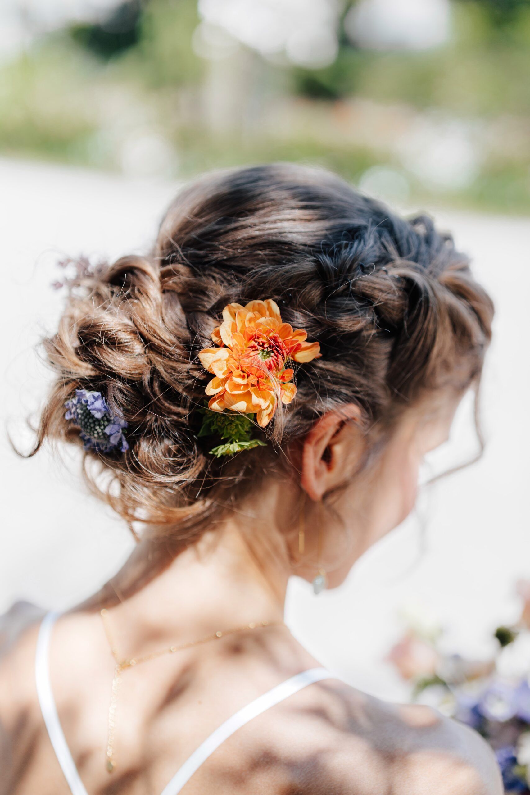 Eine Frau mit braunem, gewelltem Haar, das zu einer Hochsteckfrisur mit einer orangefarbenen Blume und kleinen lilafarbenen Blumen gestylt ist, von hinten gesehen in weichem natürlichem Licht bei einer Hochzeit bei Tante Polly Karlsruhe.