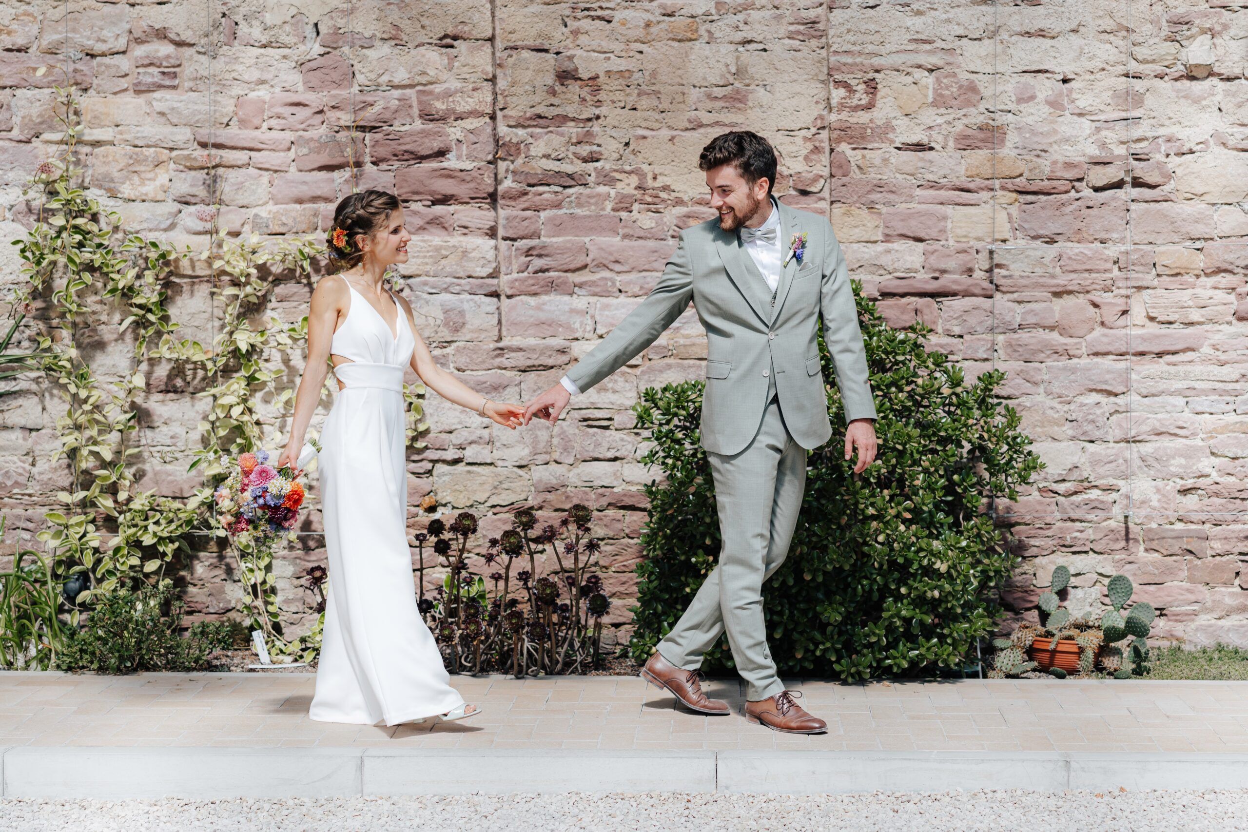 Eine Braut in einem weißen Kleid mit einem bunten Blumenstrauß geht Hand in Hand mit einem Bräutigam in einem hellgrauen Anzug lächelnd vor einer Steinmauer mit Grünpflanzen bei Hochzeit Tante Polly Karlsruhe.