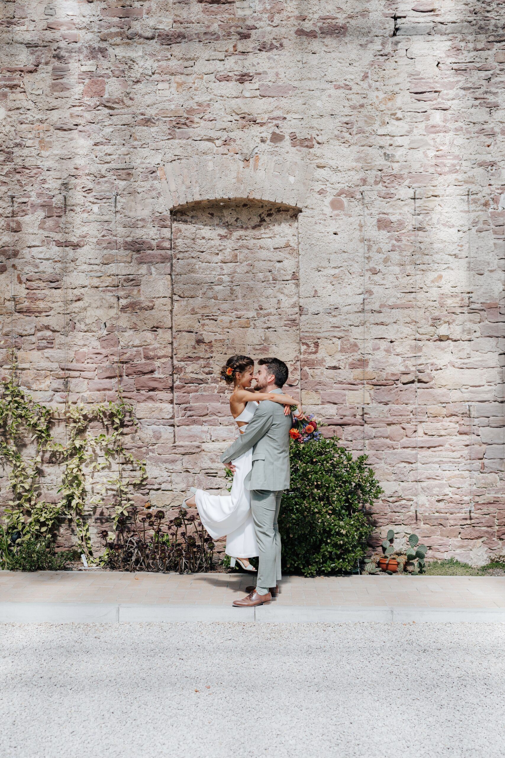 Ein Bräutigam hebt seine Braut hoch und umarmt sie vor einer rustikalen Steinmauer bei Hochzeit Tante Polly Karlsruhe. Die Braut hält einen Blumenstrauß und ihr Kleid schwingt, während sie einander anlächeln, umgeben von Grün zu ihren Füßen.
