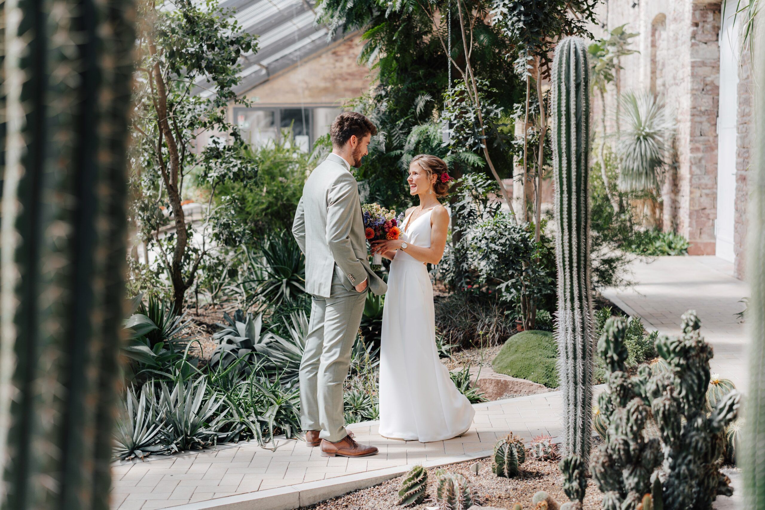 Braut und Bräutigam teilen einen zärtlichen Moment auf einem gefliesten Weg in einem üppigen Gewächshaus bei Hochzeit Tante Polly Karlsruhe, umgeben von Kakteen und Grünzeug, mit dem Bräutigam in einem hellen Anzug und der Braut, die einen Blumenstrauß hält.