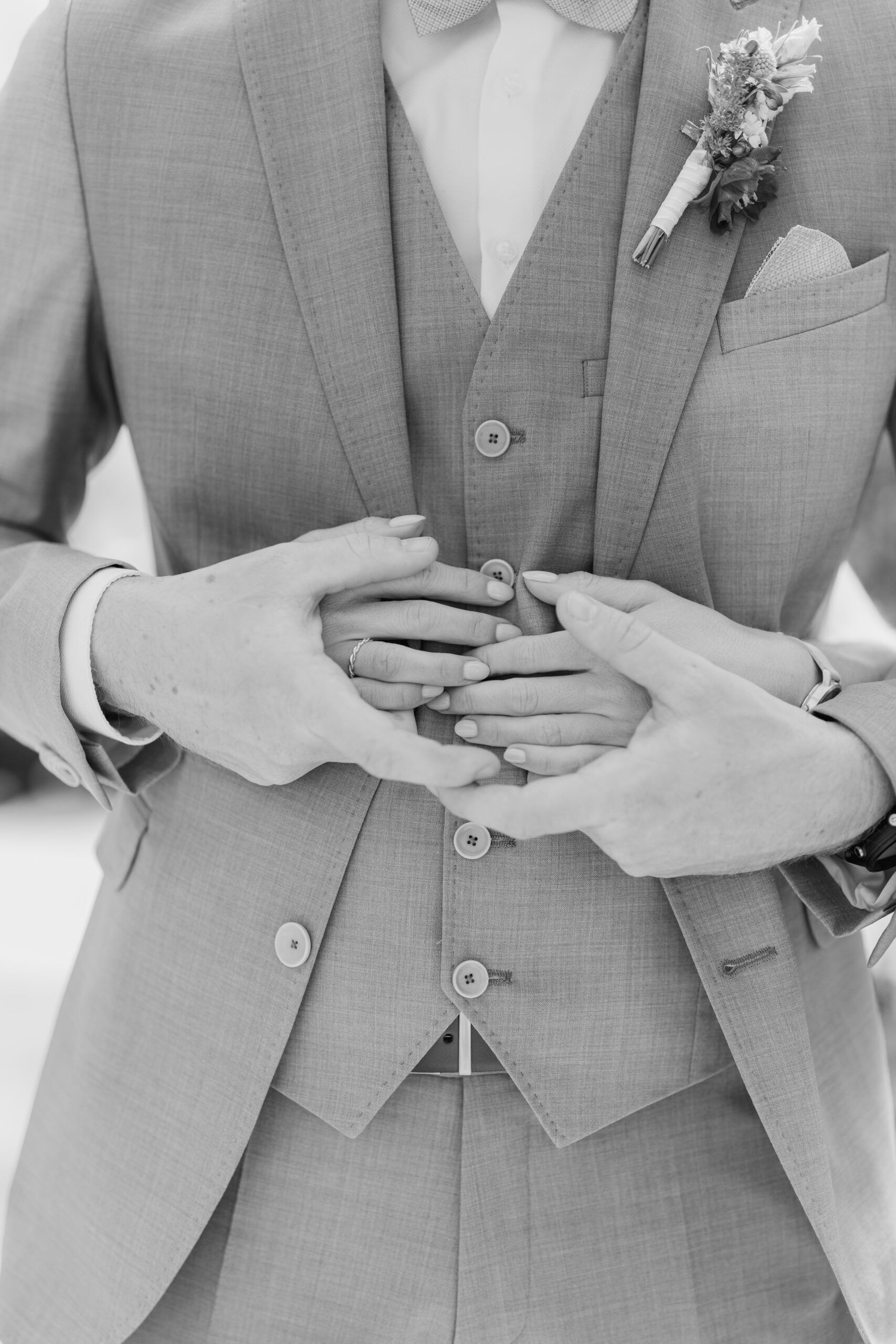 Ein Schwarz-Weiß-Foto in Nahaufnahme einer Person im Anzug mit einer Boutonnière, die sich von hinten umarmt - vier Hände mit sanft aufliegenden Eheringen, die Intimität und Verbundenheit bei einer Hochzeit bei Tante Polly Karlsruhe symbolisieren.