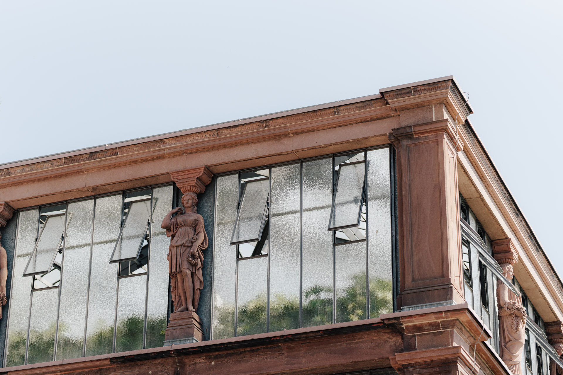 In der Ecke eines Gebäudes mit großen Milchglasfenstern, die teilweise geöffnet sind, stehen zwischen Säulen klassische Statuen von Frauen in Gewändern, die an eine Hochzeit bei Tante Polly in Karlsruhe erinnern.